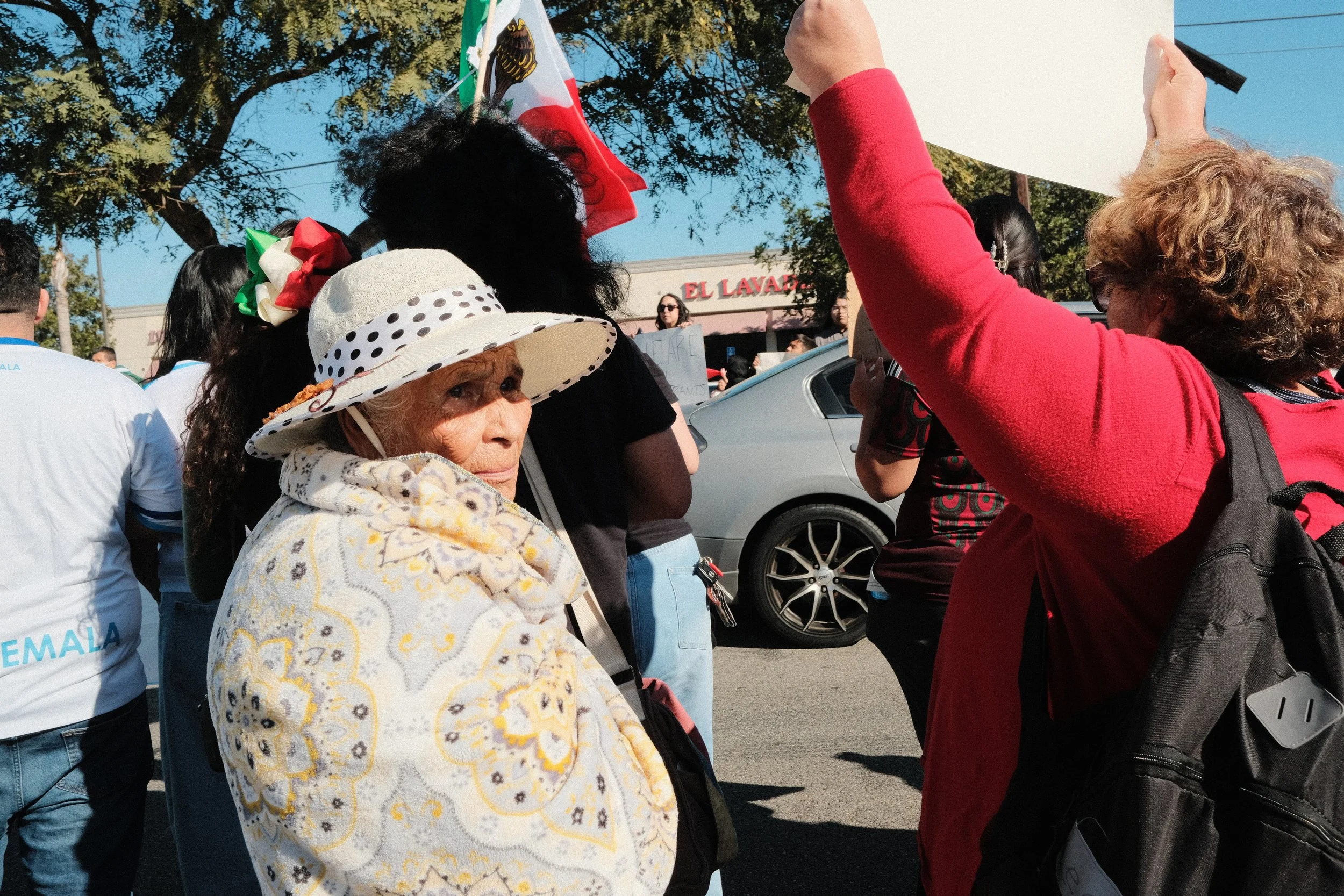 Group of people gathered outdoors during a demonstration or rally, with some holding signs and flags, including a Mexican flag, and wearing casual clothing.