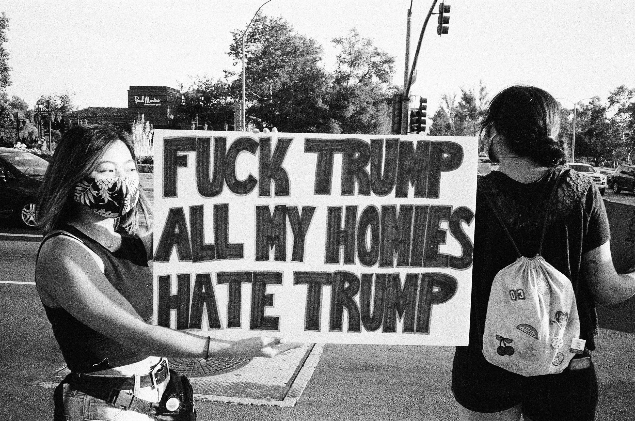 Two young women holding a protest sign that reads, 'F*** TRUMP ALL EYES ON HATE TRUMP.' One woman is wearing a floral face mask, and the other has a backpack with cherry and other patches. They are standing on a street with cars and traffic lights in