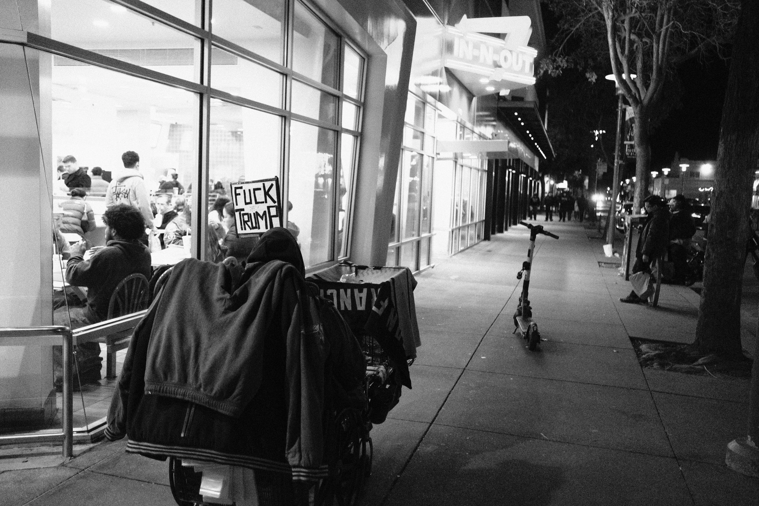 Night scene outside a fast food restaurant with people sitting inside, visible through large windows. In the foreground, a sign with offensive language is placed outside. To the right, pedestrians and parked bikes line the sidewalk, and there are tre
