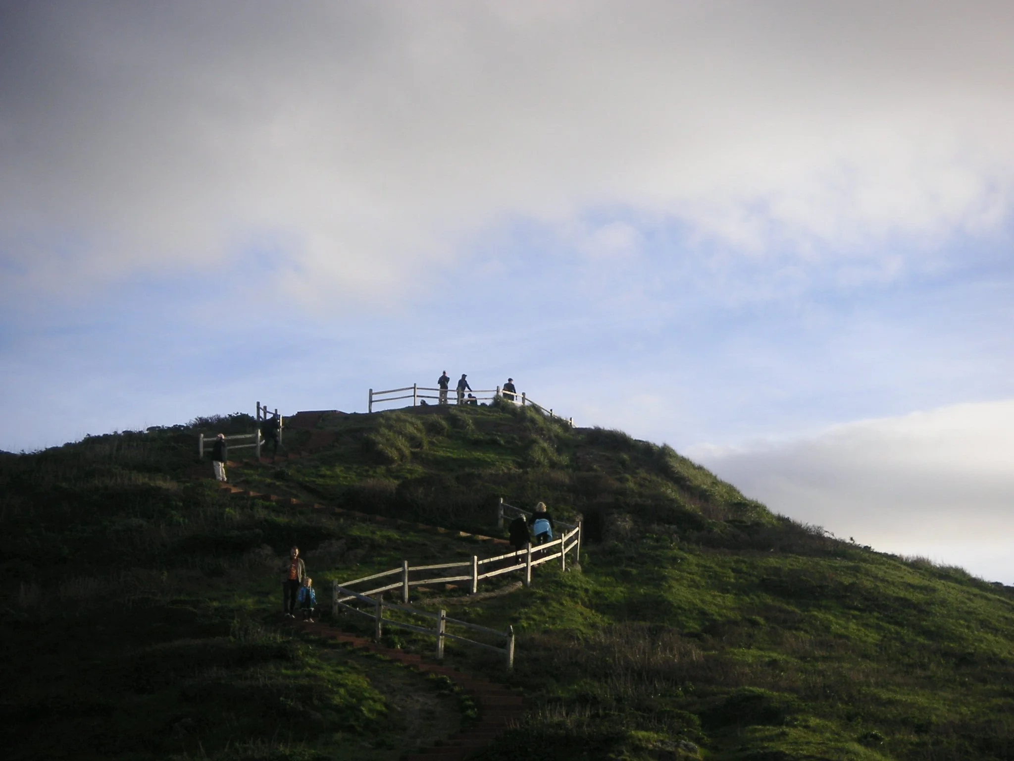 People walking up a hill with a fence and pathway, under a cloudy sky.