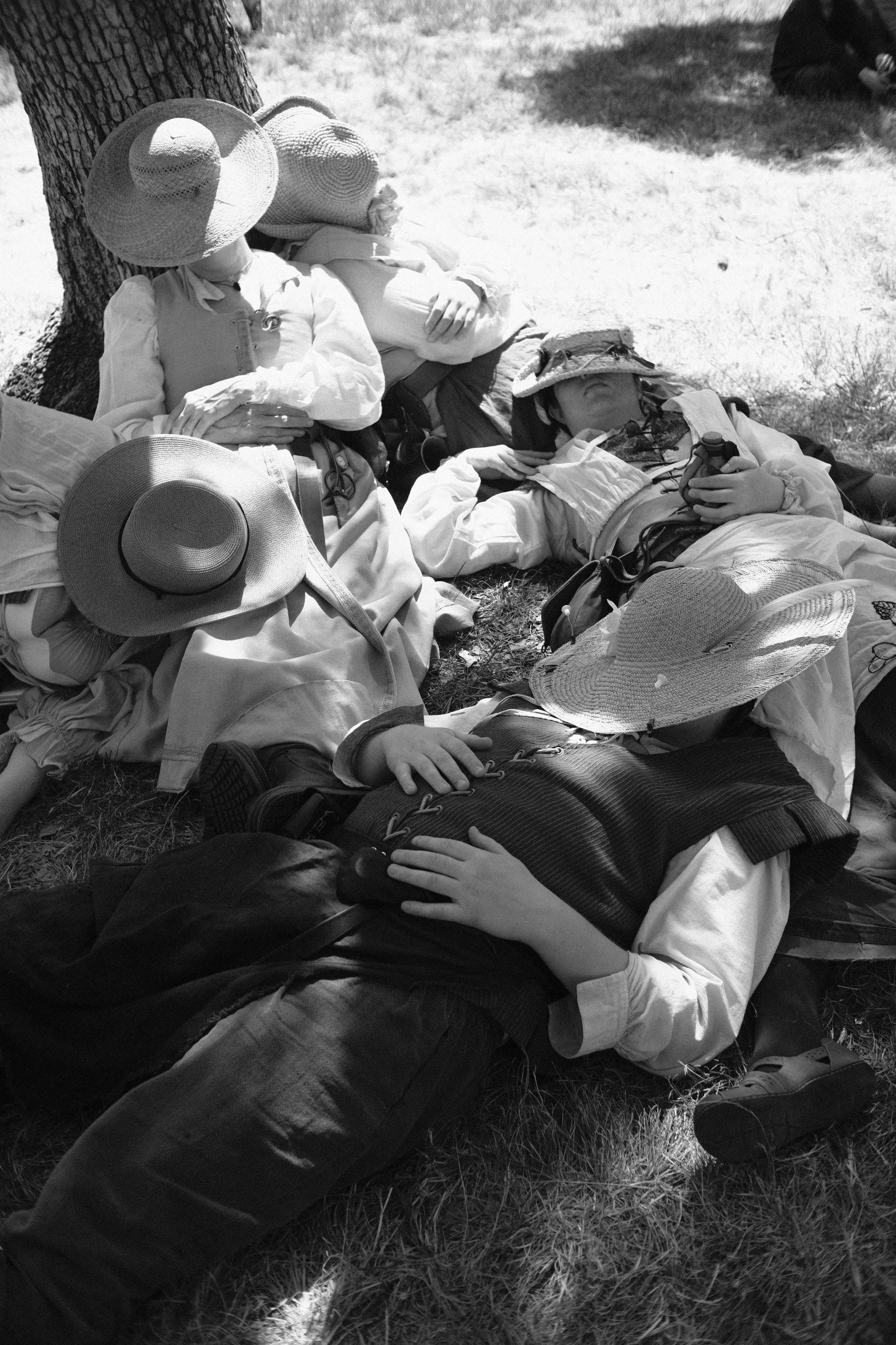 Four people are resting on the grass outdoors, two are lying down with wide-brimmed hats covering their faces, and two are sitting under a tree, one is leaning against the trunk and the other has their hands crossed in their lap. They are dressed in 