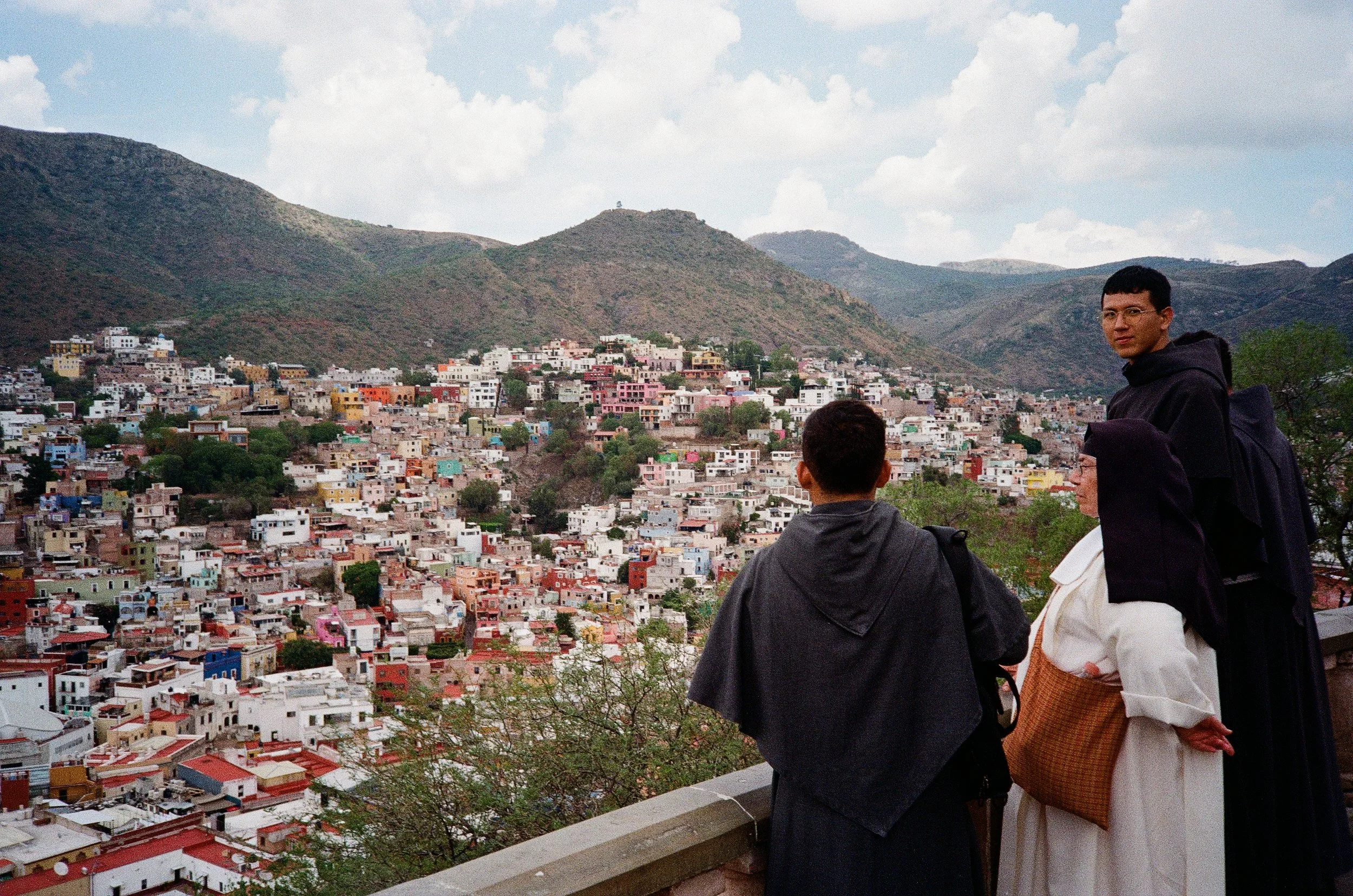 Three people standing on a balcony overlooking a colorful hillside neighborhood with mountains in the background.
