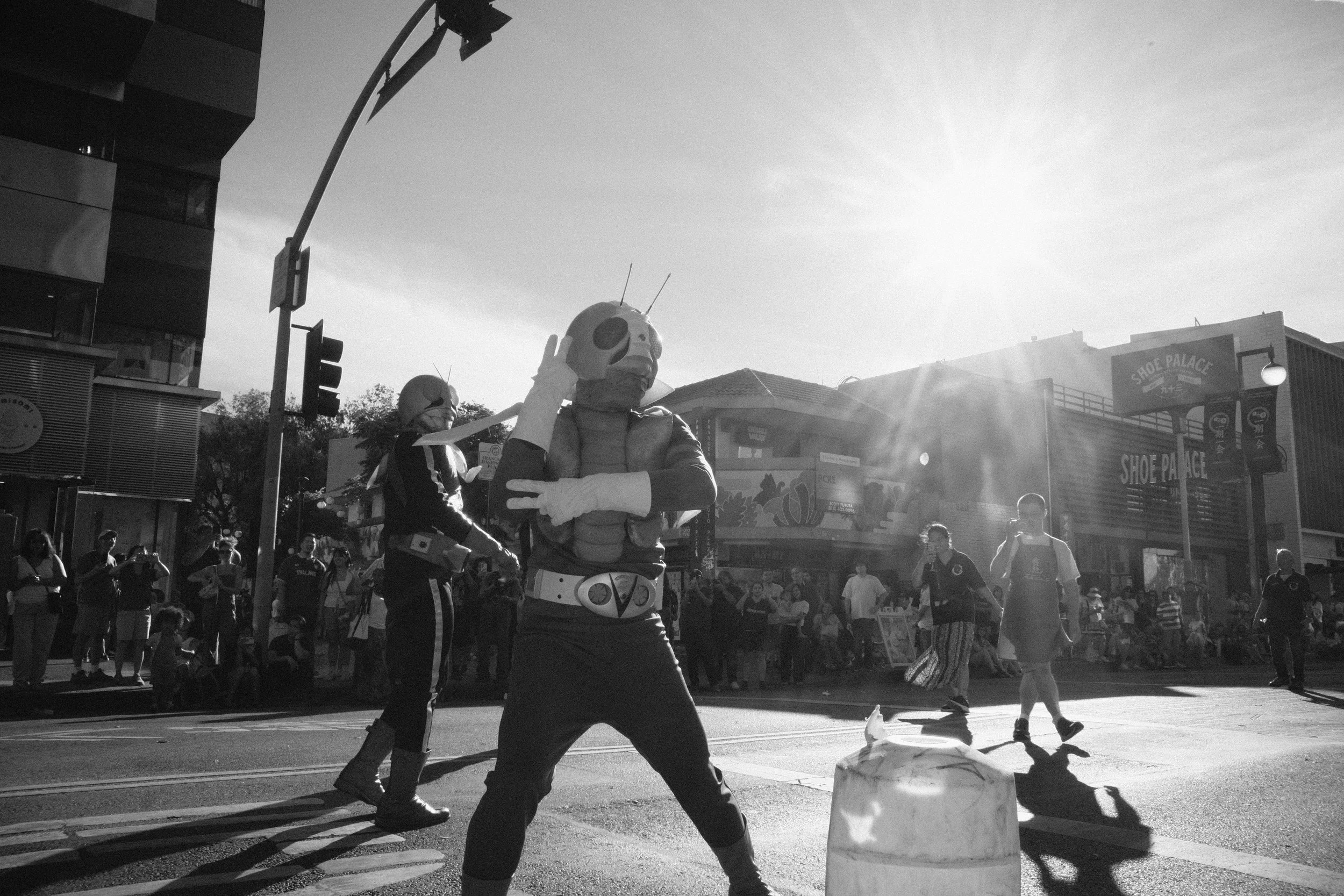Street scene with a person dressed as a skeleton in a superhero costume, and another person in a warrior costume, among a crowd of onlookers. The scene is in black and white, with sunlight creating lens flare.