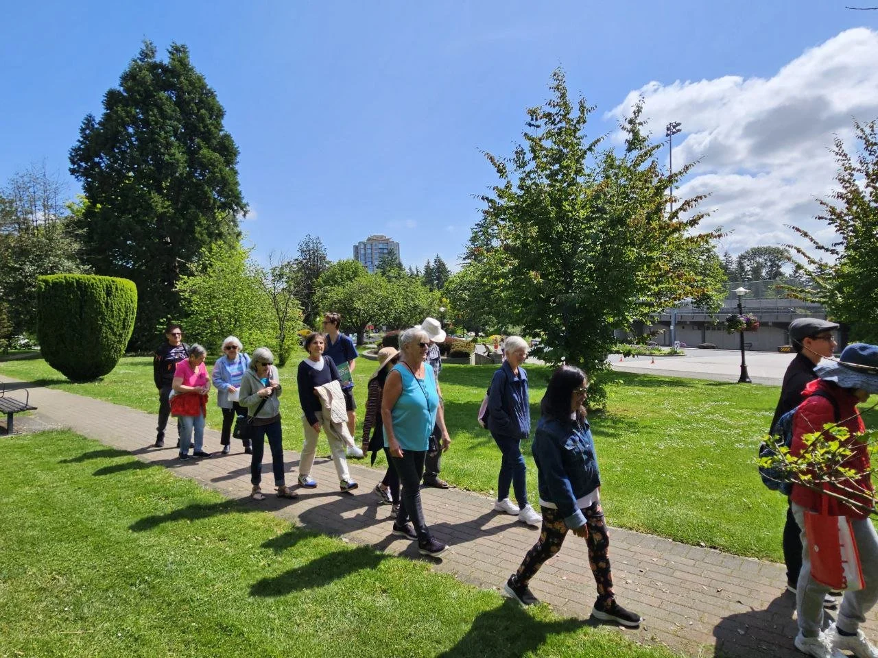 A group of older adults walking together along a paved path through a sunny, tree-lined park