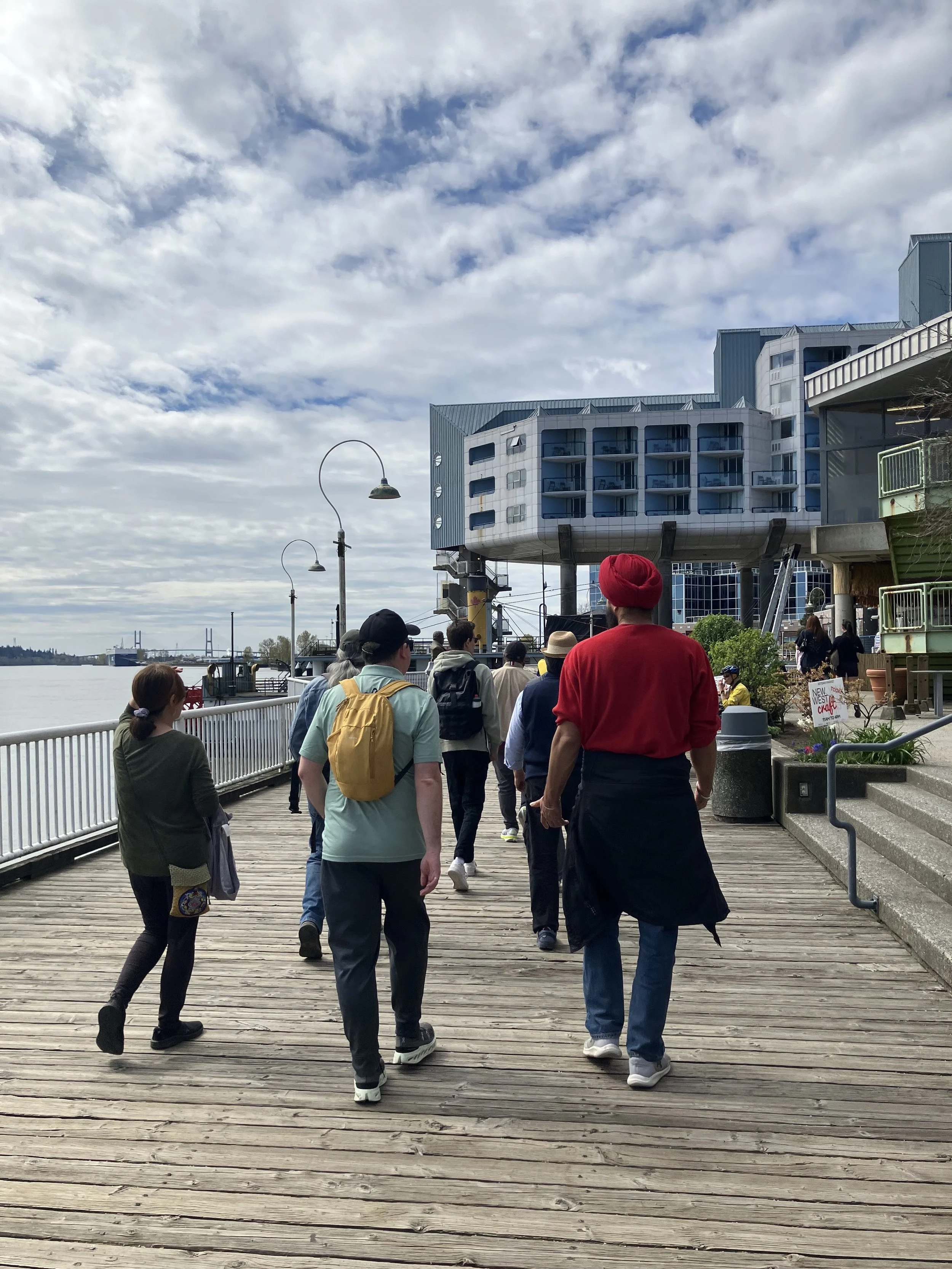 A group walks along a wooden riverfront boardwalk past shops and a hotel, with the river and a bridge in the distance