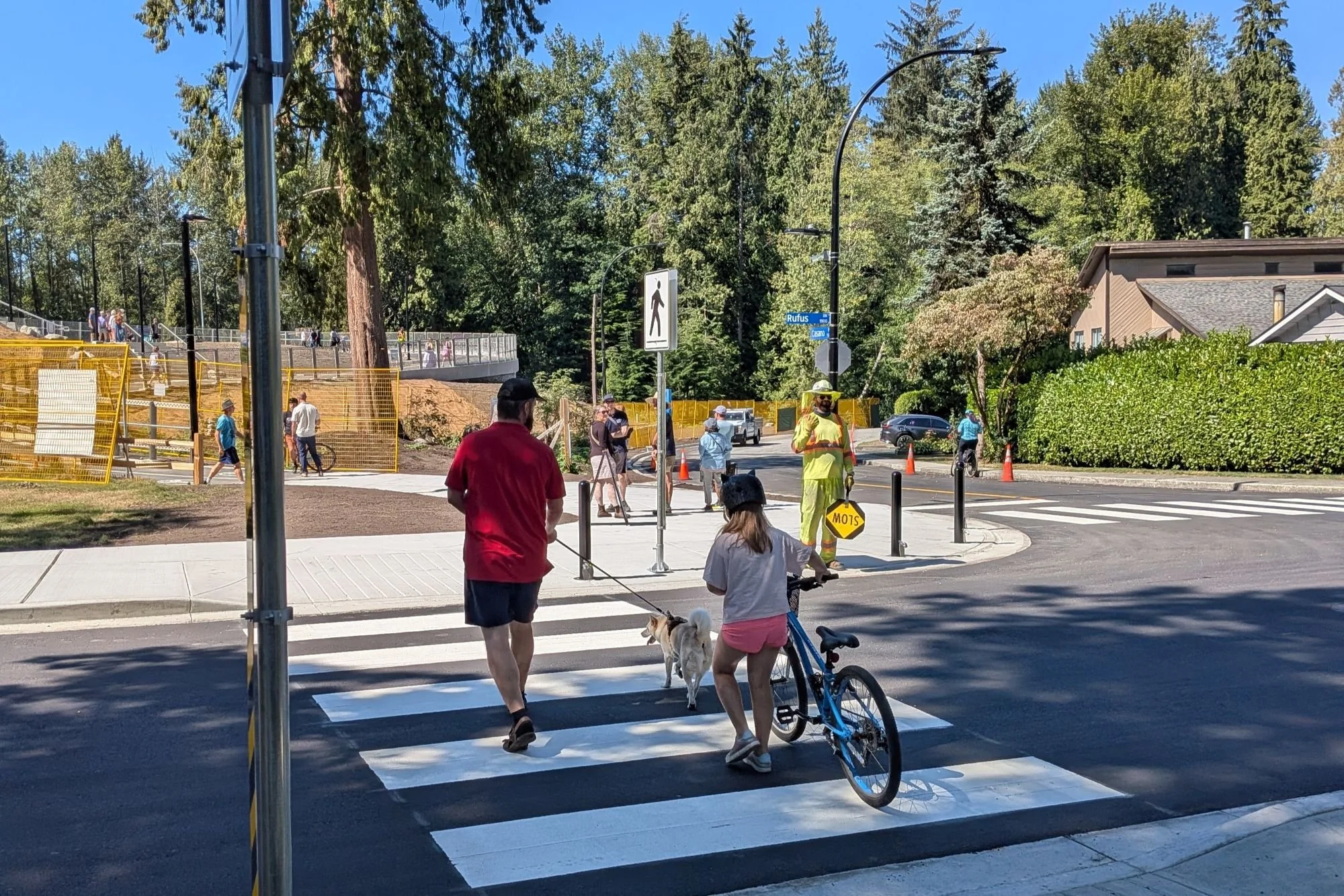 A parent walking a dog and a child walking a bike cross a wide, freshly painted crosswalk while a crossing guard watches