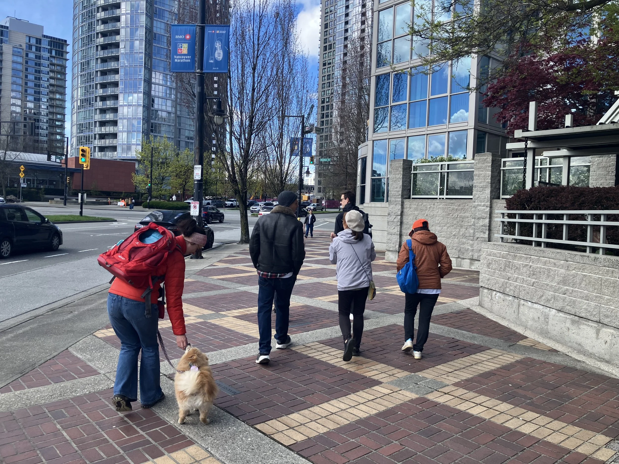 A small group walks along a wide, level brick sidewalk downtown, one person leaning down to a small fluffy dog on a leash.