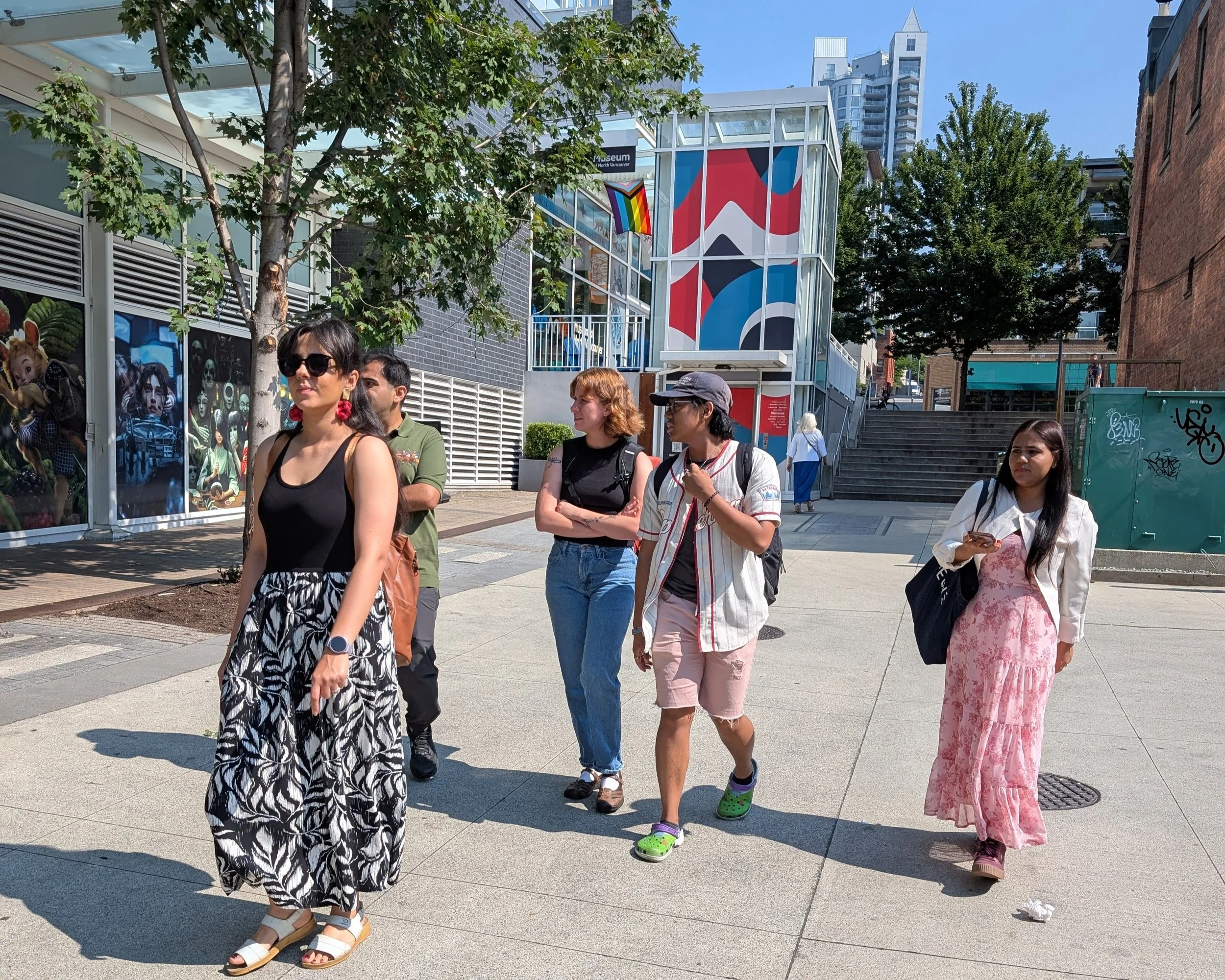 A group of young adults walks across a sunny public plaza past colourful murals and street trees.