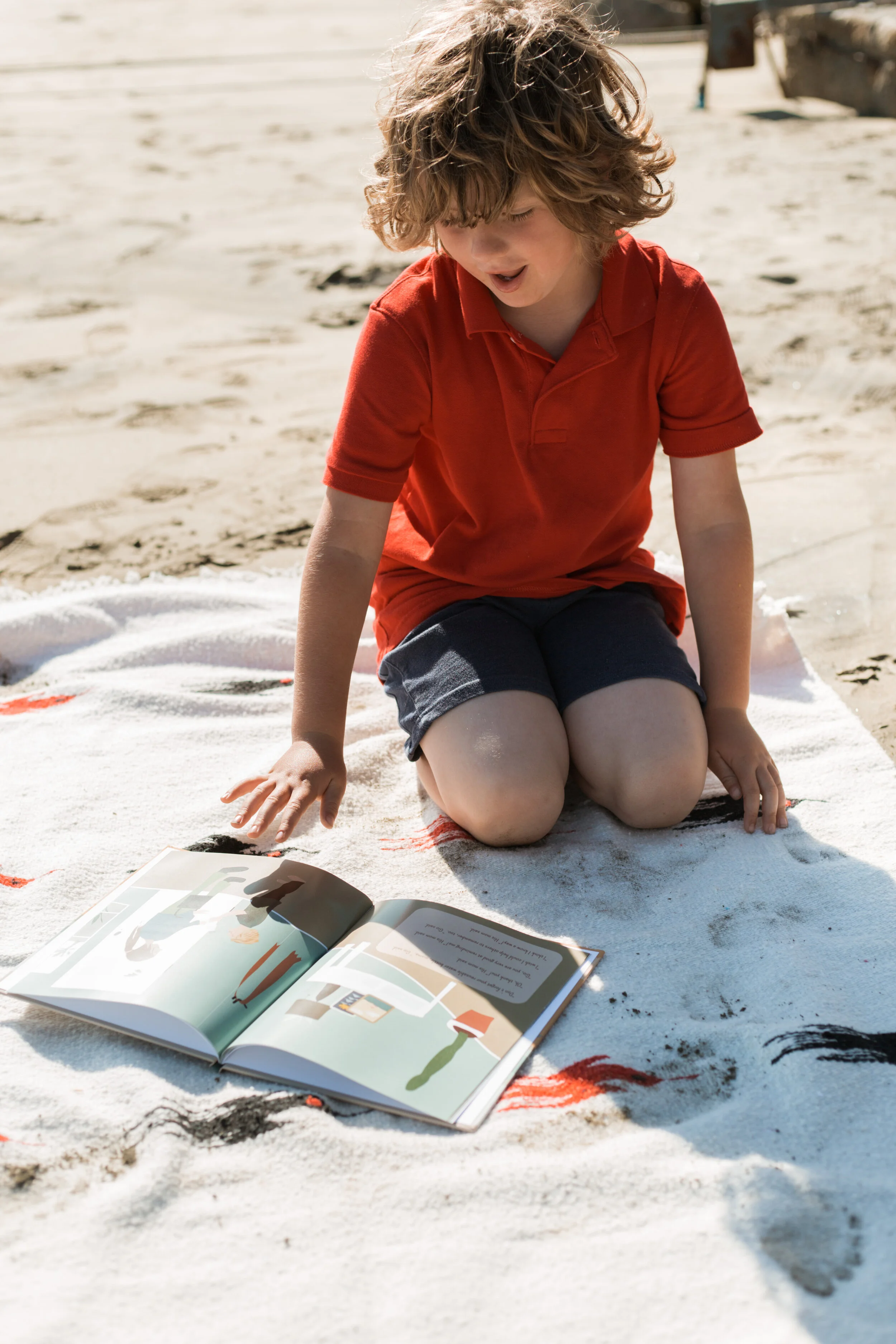 Young boy in a red shirt sitting on a sandy beach, reading a picture book on a white blanket.