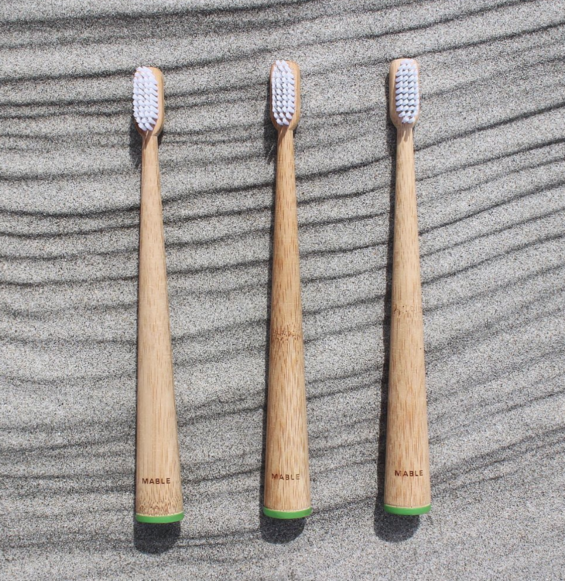 Three MABLE eco-friendly toothbrushes with green handles resting on a sandy surface, promoting sustainable dental care.