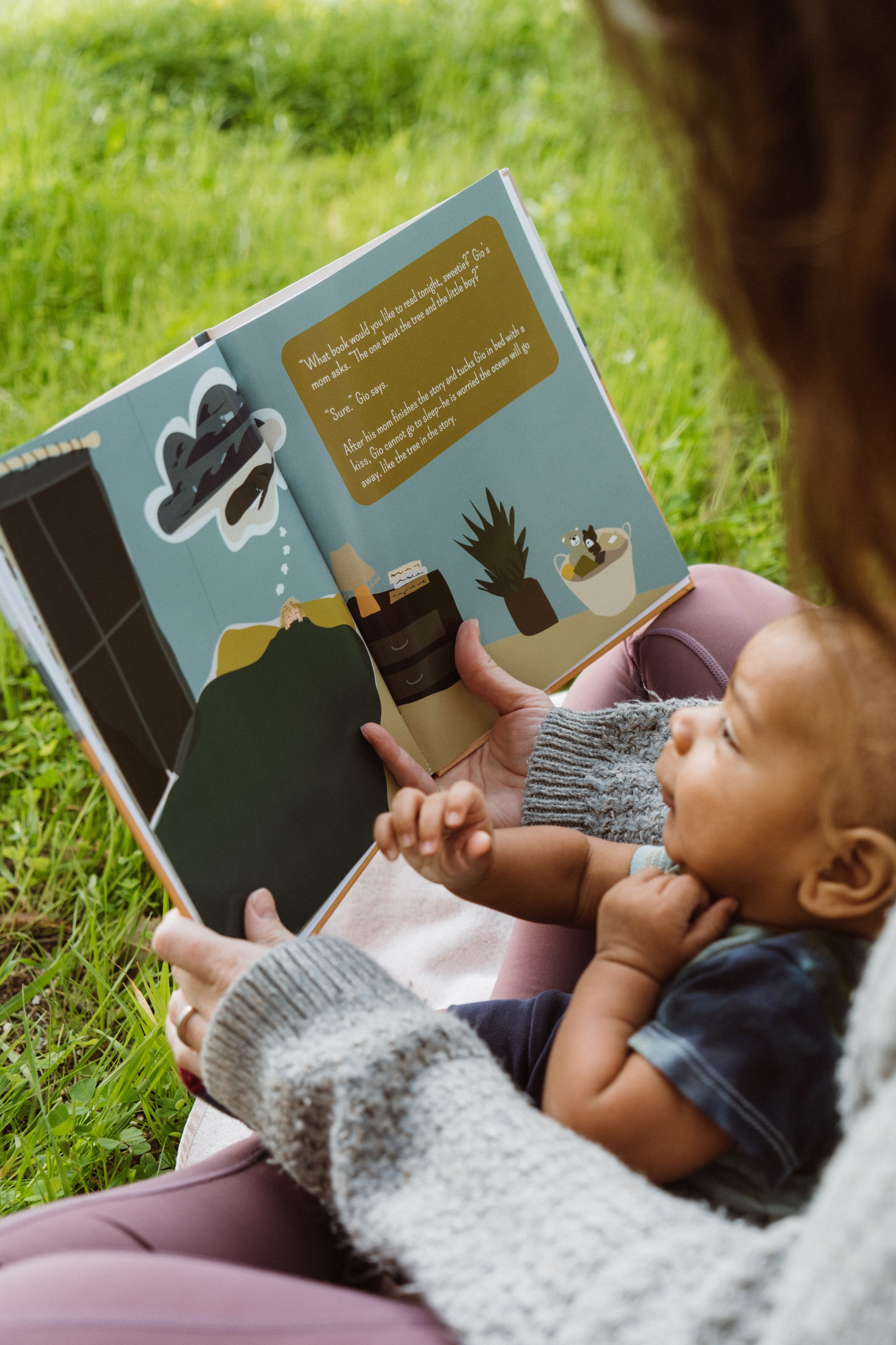 Close-up of a mother reading a picture book to her attentive baby while sitting on a blanket in a green field.