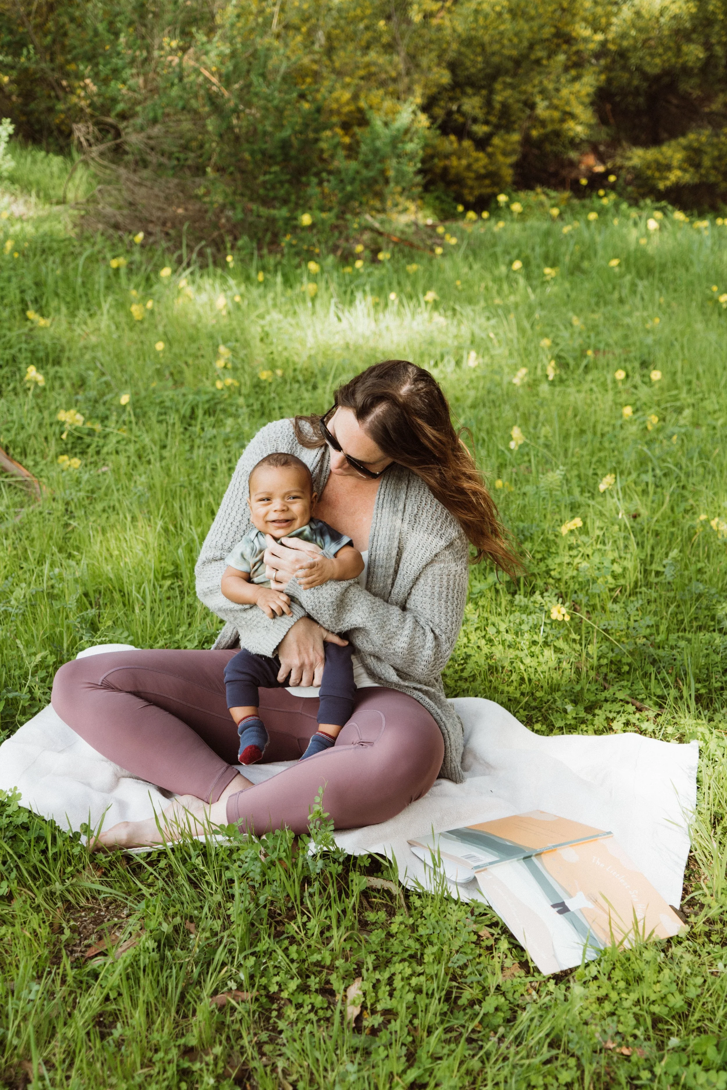 Mother sitting on a blanket in a lush green field, holding her smiling baby and reading a book.