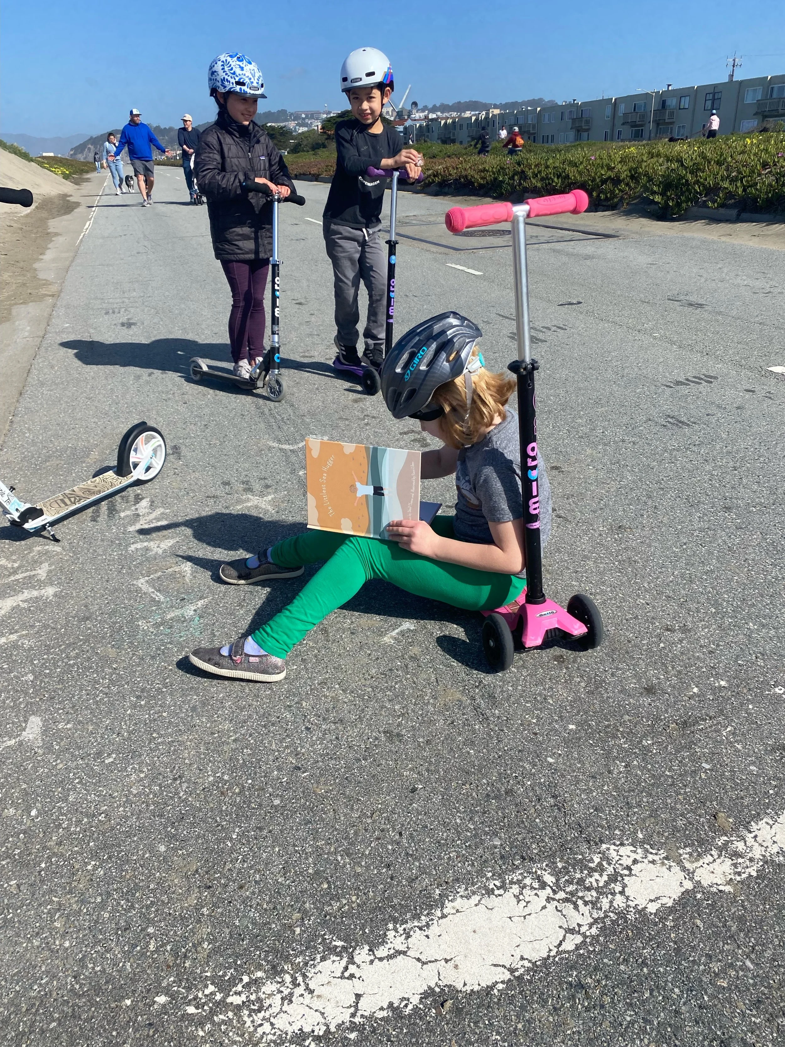 Children with scooters enjoying a sunny day outdoors, one child sitting on the pavement reading a book