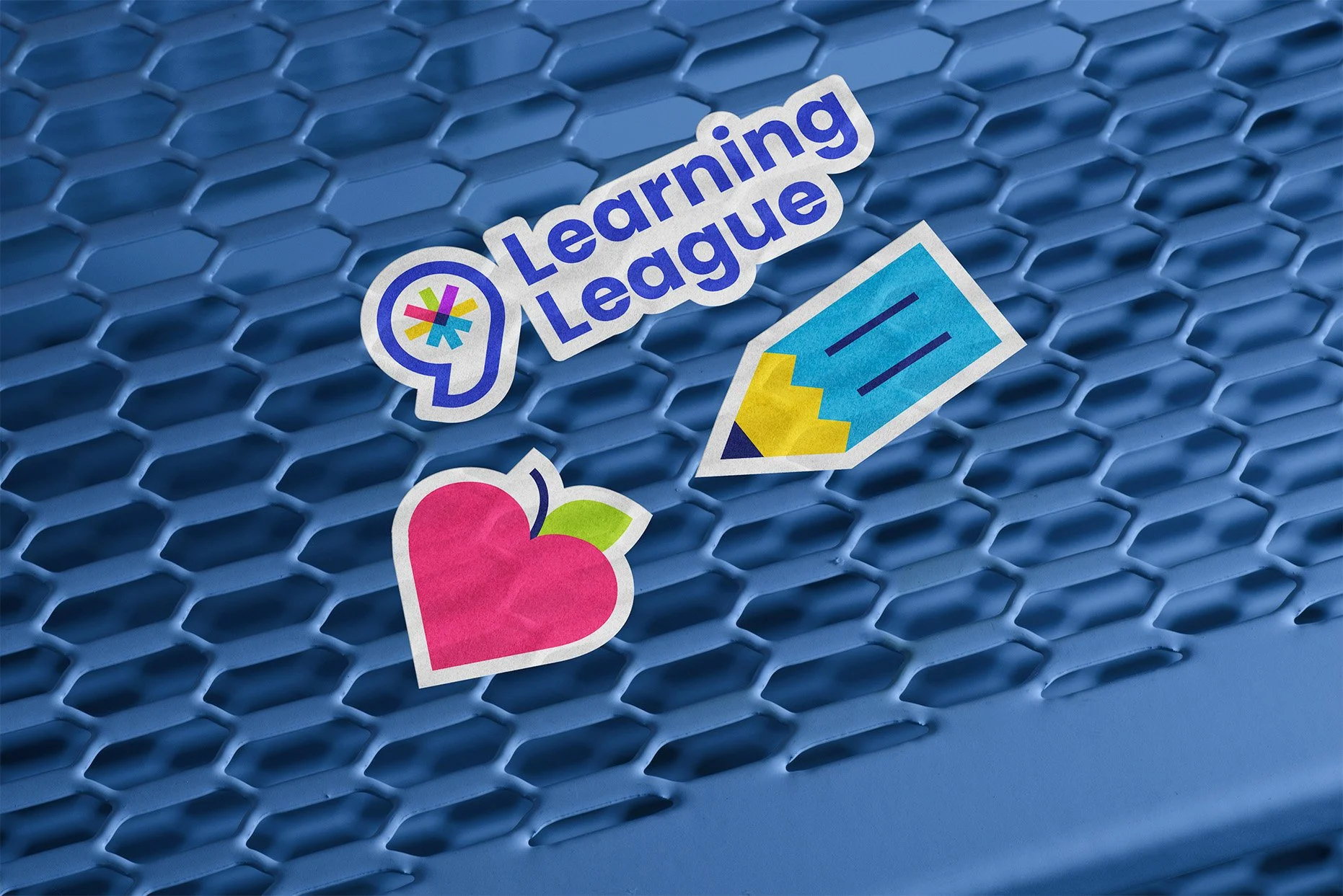 Three stickers are stuck to an outdoor table. One is the Learning League logo, one is a pencil, and one is a heart-shaped apple