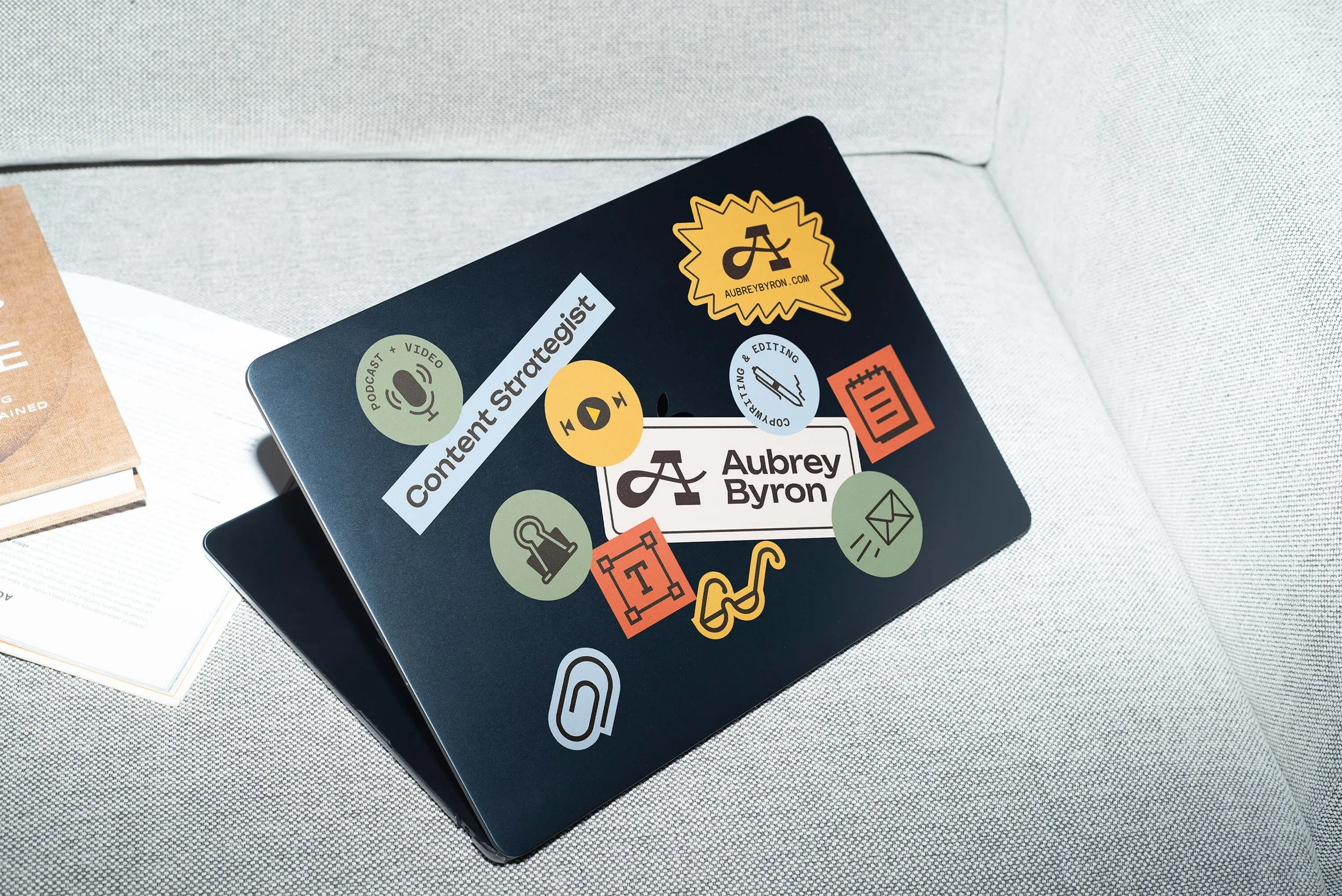 A Macbook laptop covered in Aubrey Byron branded stickers sits next to books on a couch