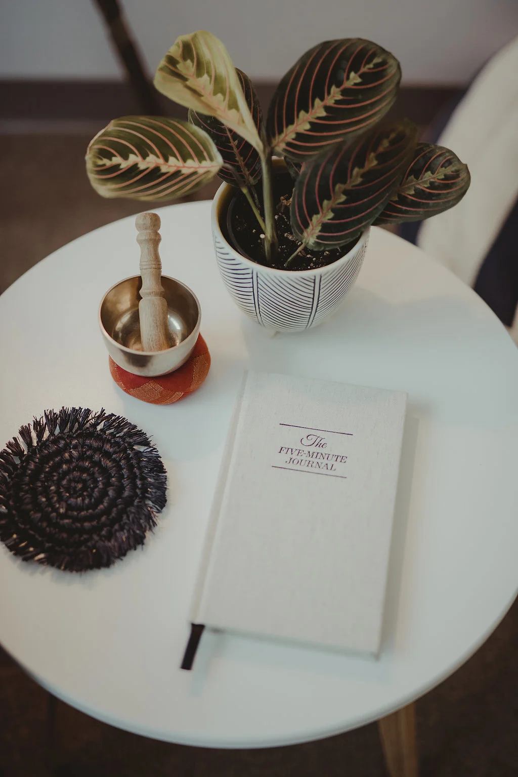 Image of a small round white table. On top of the table there is a small houseplant, a meditation bowl sitting on a red and gold cushion, a woven dark blue drink coaster, and a white journal that says "The Five Minute Journal" on the cover.