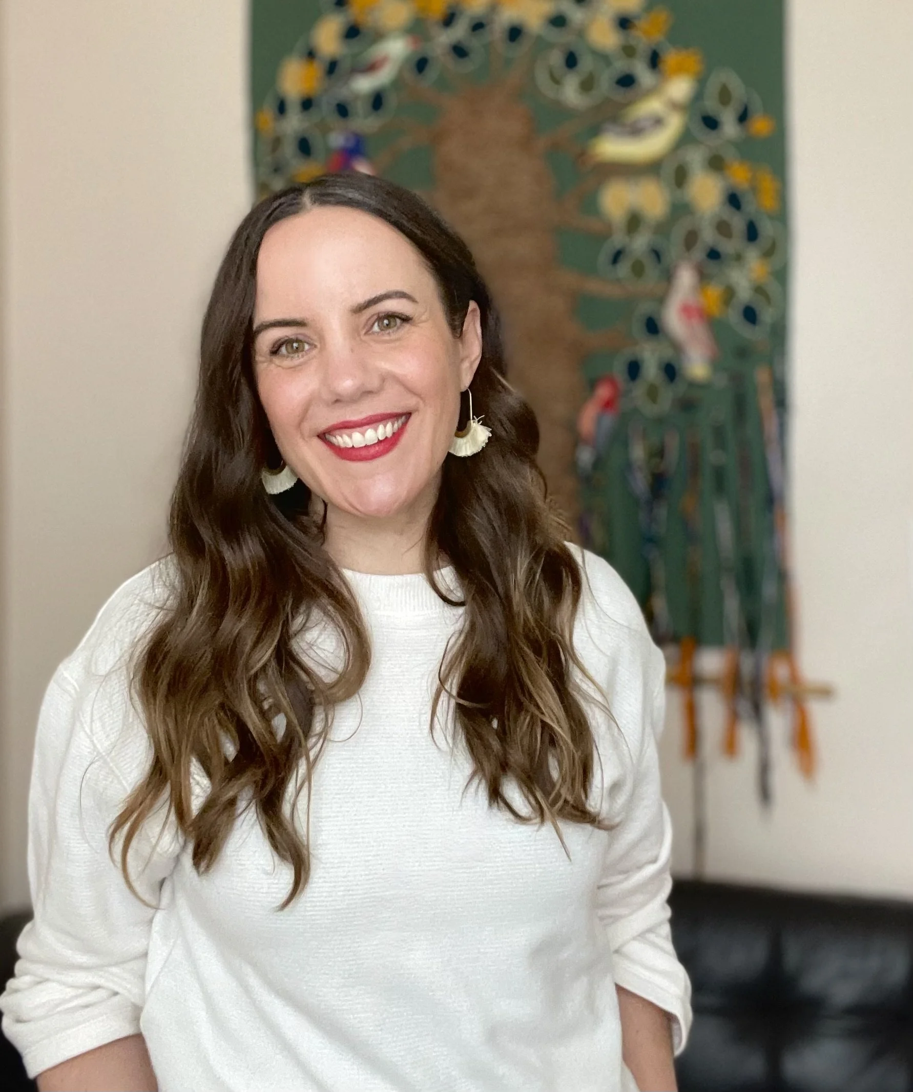Image of therapist with long brown hair and hazel eyes, wearing a white top, standing in a room in front of a green wall tapestry. She is smiling at the camera.