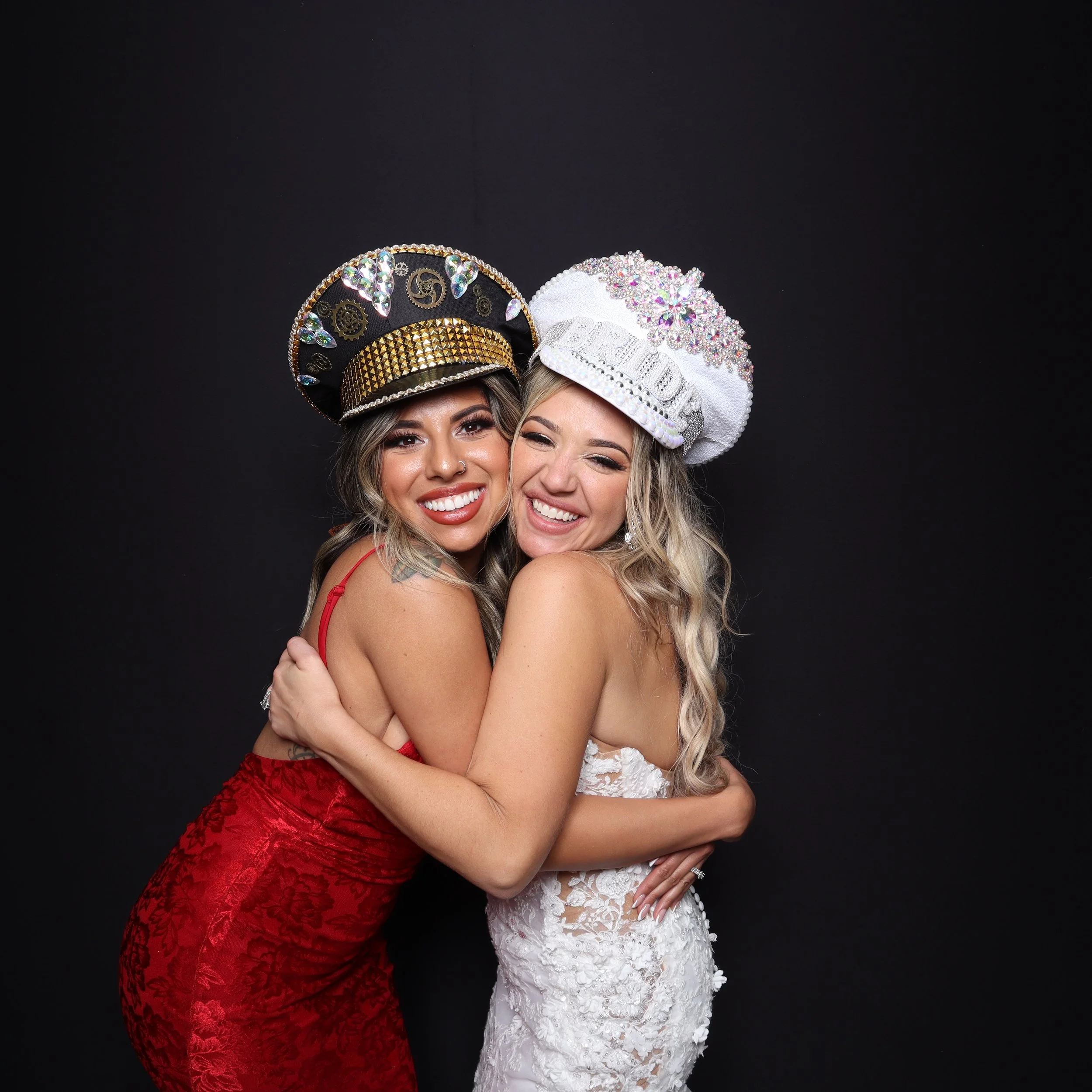 Two women wearing decorative hats, embracing and smiling against a black background. One is in a red dress, the other in a white lace dress.