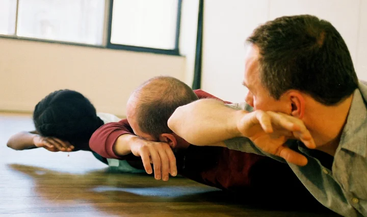 Three people lying face down on a wooden floor, stretching or doing a yoga pose, with their arms extended forward.