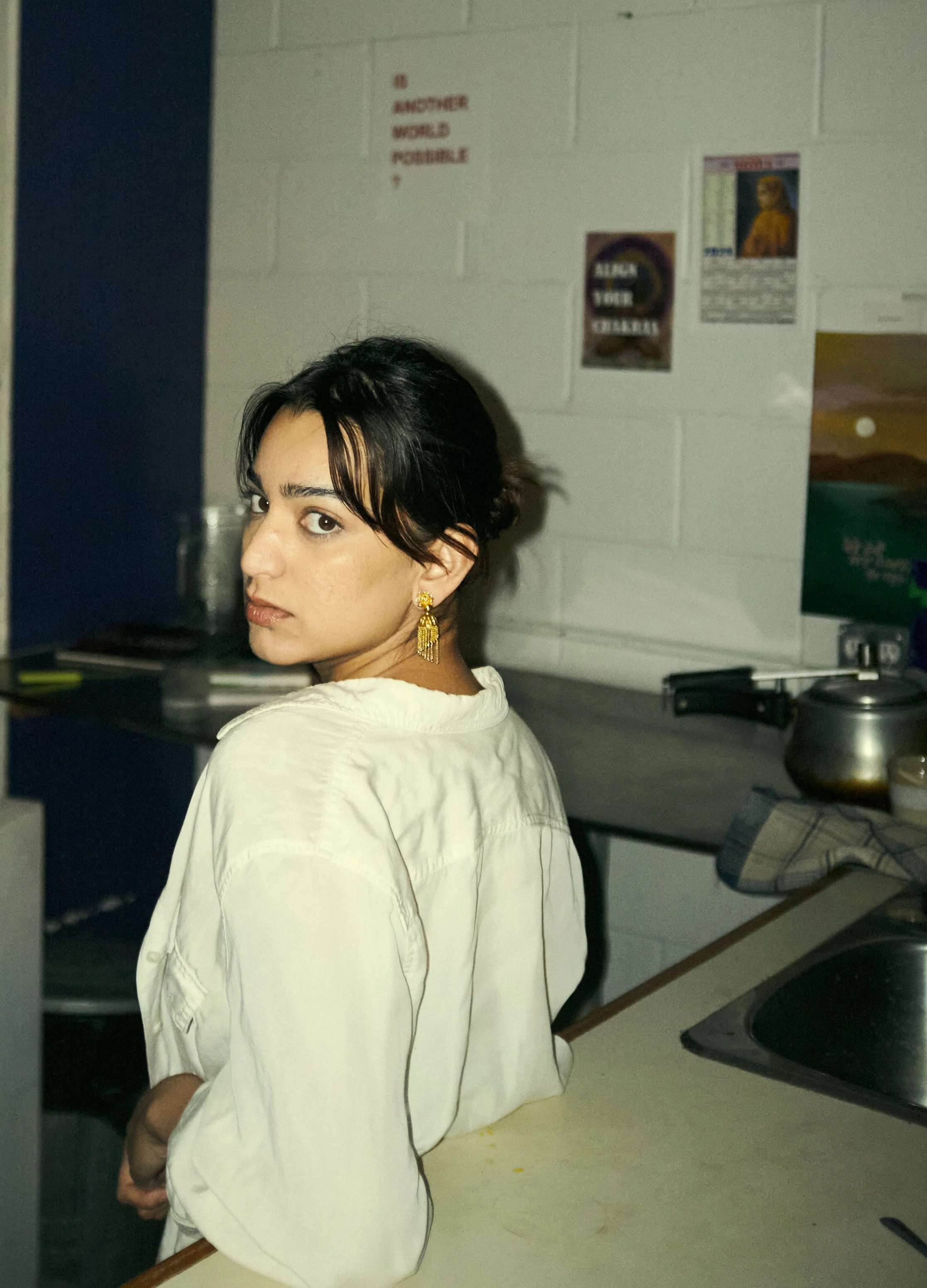 A woman with short black hair and gold earrings looking over her shoulder at the camera in a kitchen or break room with posters on the wall and a metal pot on the counter.