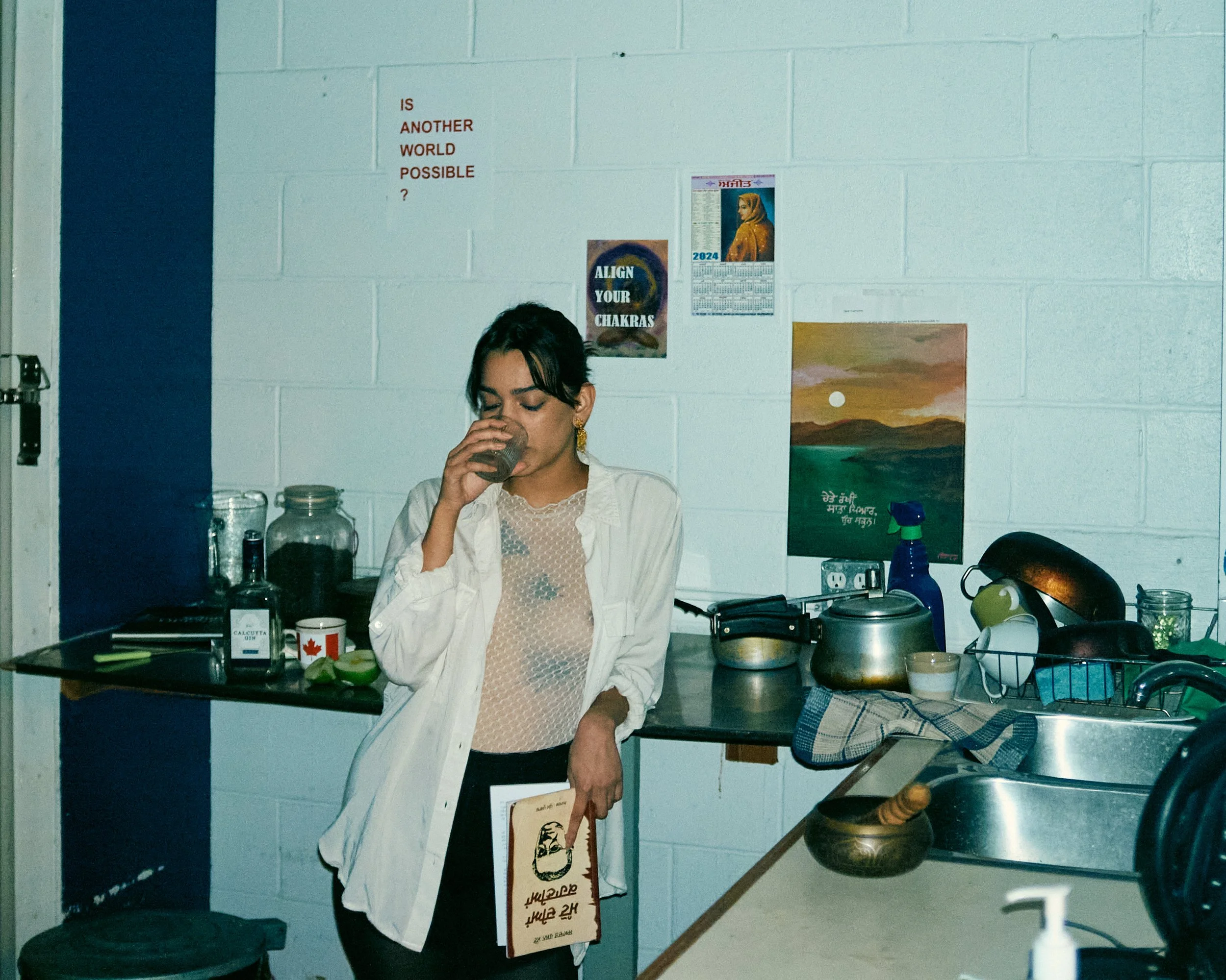 A woman with dark hair in a bun drinks from a glass in a kitchen. There are various kitchen items such as jars, a cutting board, and a teapot on the counter. The wall behind her has posters and a calendar. A woman is partially visible on the right si