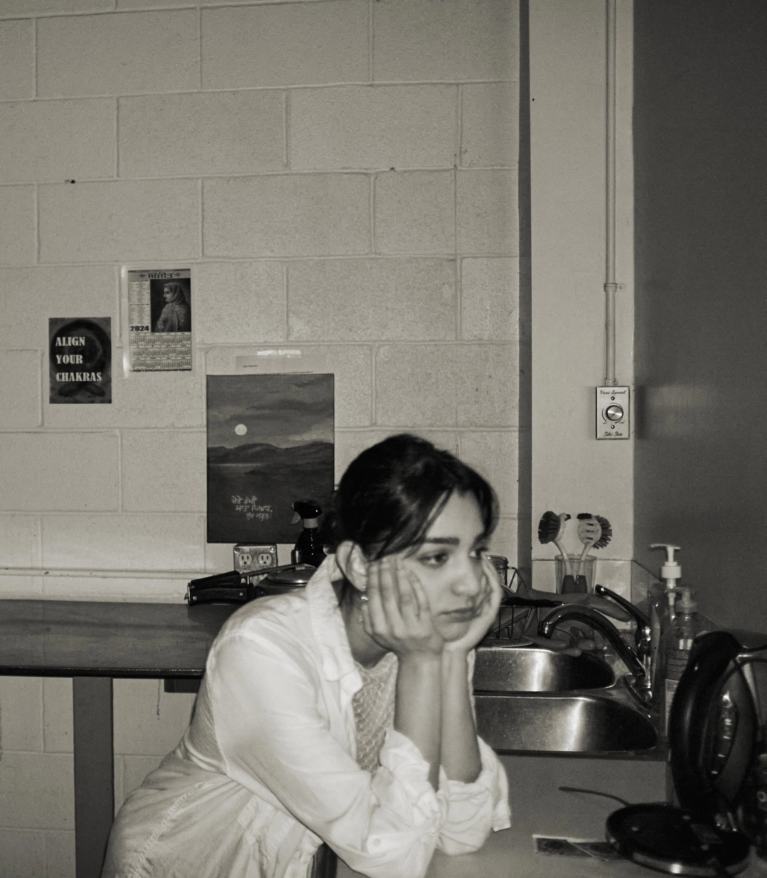 A woman sitting at a sink in a kitchen, wearing a checkered face mask, with her head bowed and hands covering her face. The background shows a gray brick wall with a poster and a calendar, and various kitchen items including a dish soap dispenser, sc