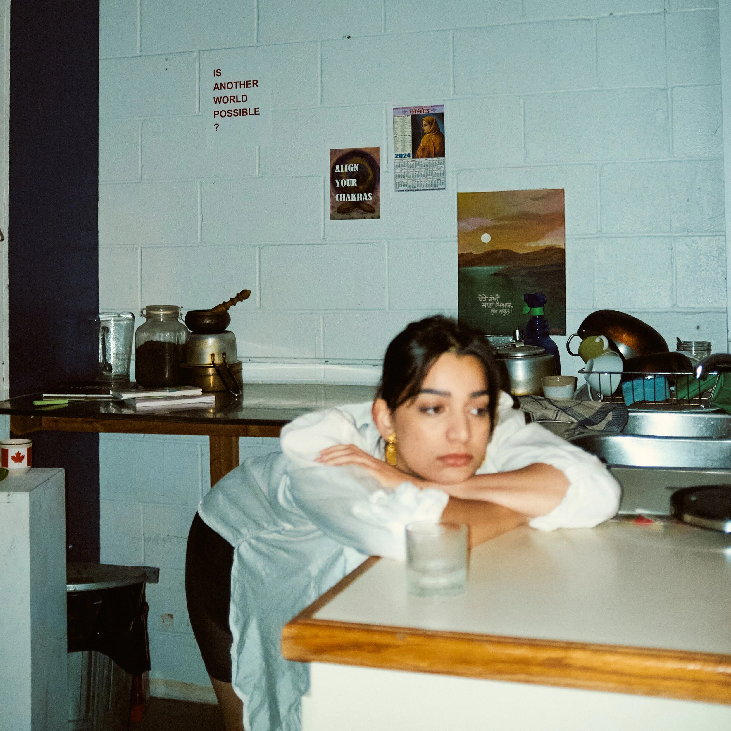A woman with dark hair and earrings resting her head on her arms on a kitchen counter, with various jars and kitchen items behind her and a wall decorated with posters and a calendar.