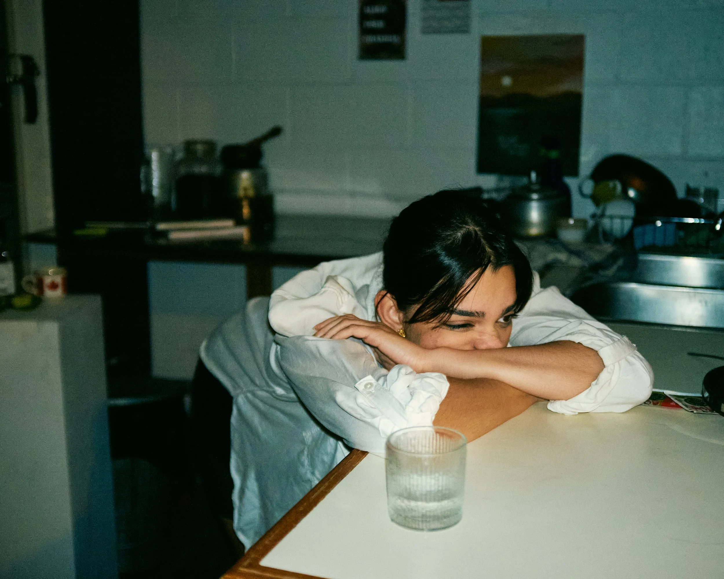 A woman with dark hair, wearing a white shirt, resting her head on her crossed arms on a table, smiling and winking. The background shows a kitchen with various items and a computer monitor.
