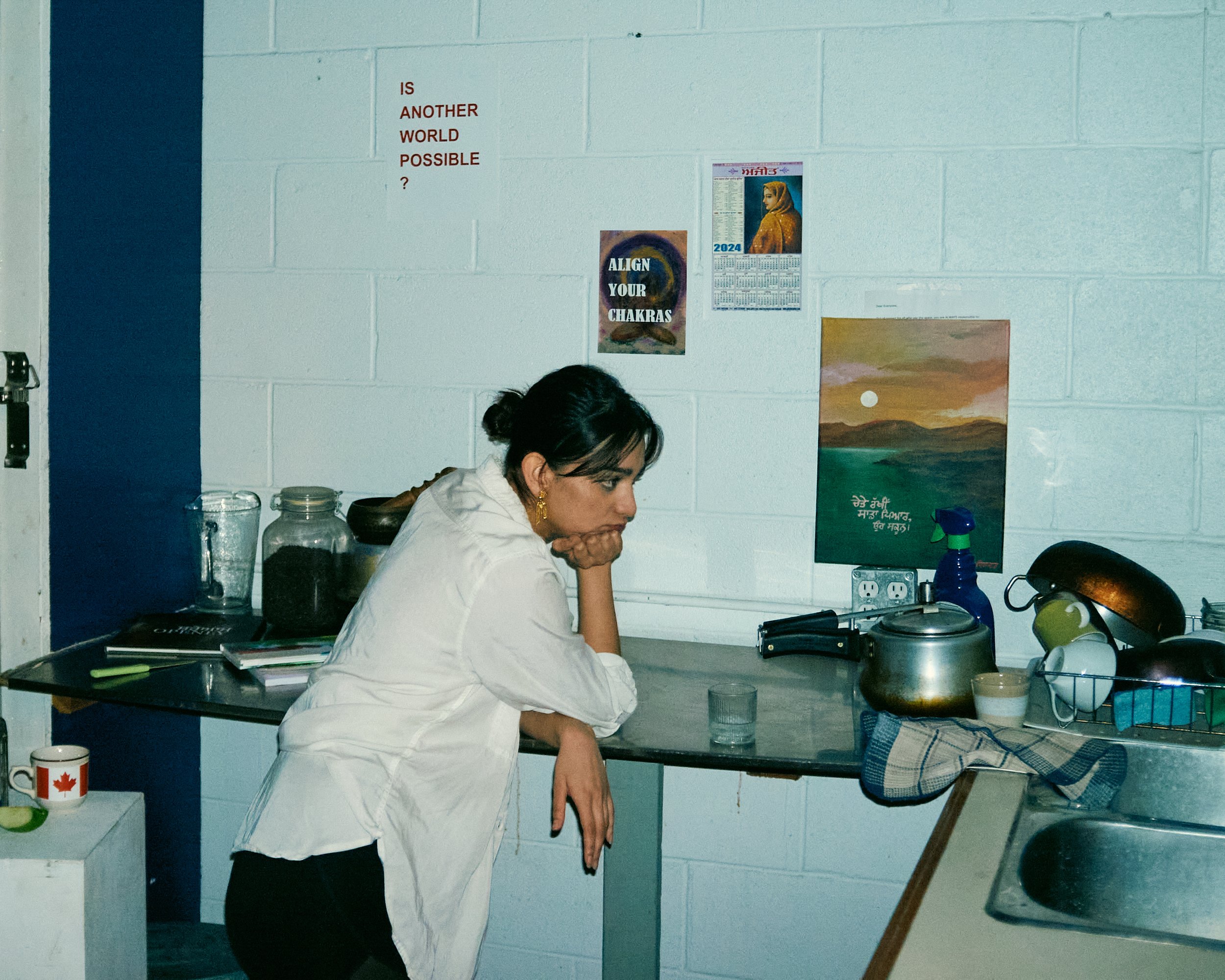 A woman is sitting at a kitchen counter with her head resting on her hand, wearing glasses and earrings, in a room with white brick walls decorated with various posters and a calendar. The counter has jars, a mug, and kitchen items.