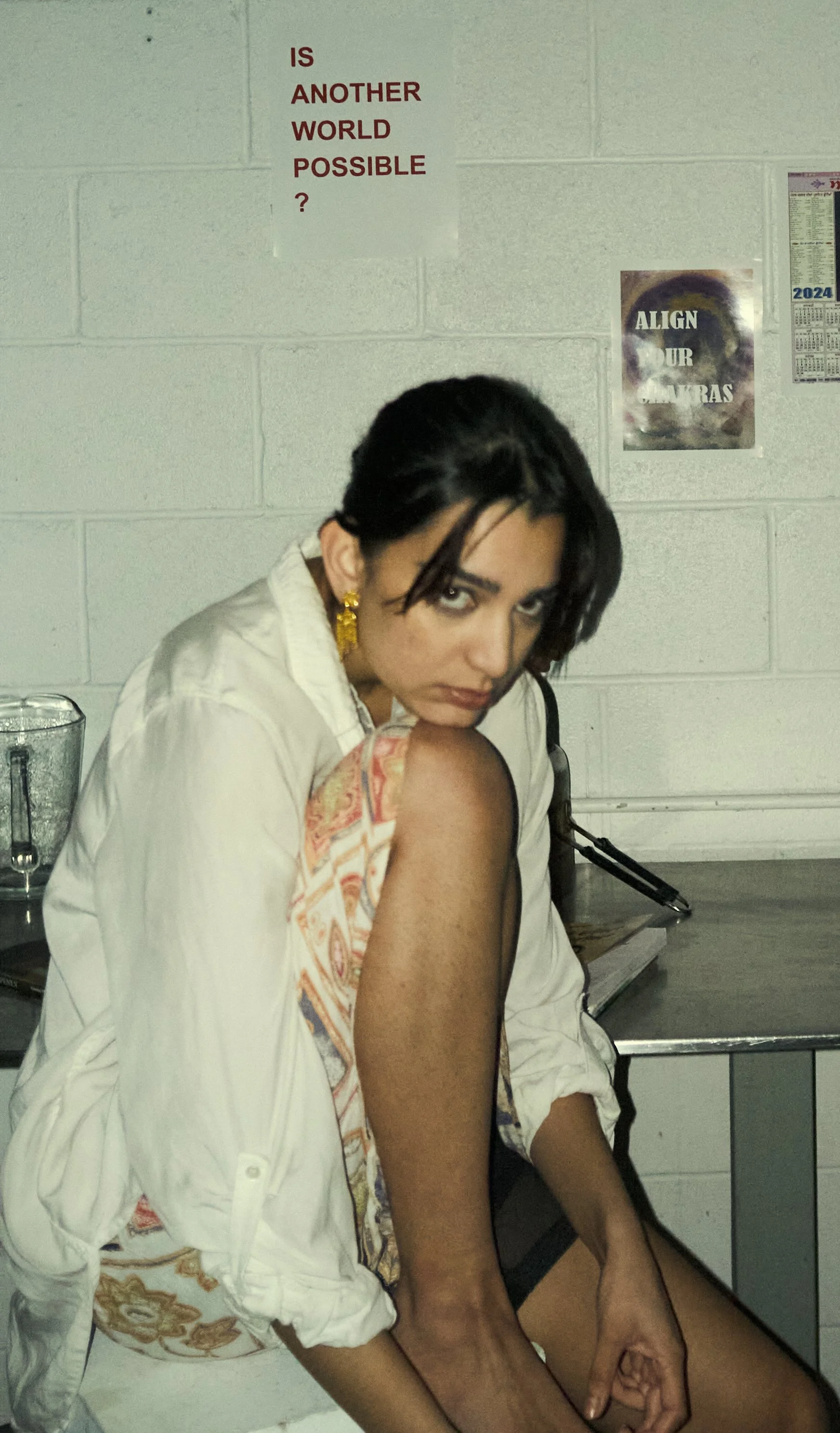 A young woman with dark hair and gold earrings sitting at a table, looking at the camera with a serious expression. Behind her on a white brick wall are posters, one with text about making another world possible and another about aligning your chakra