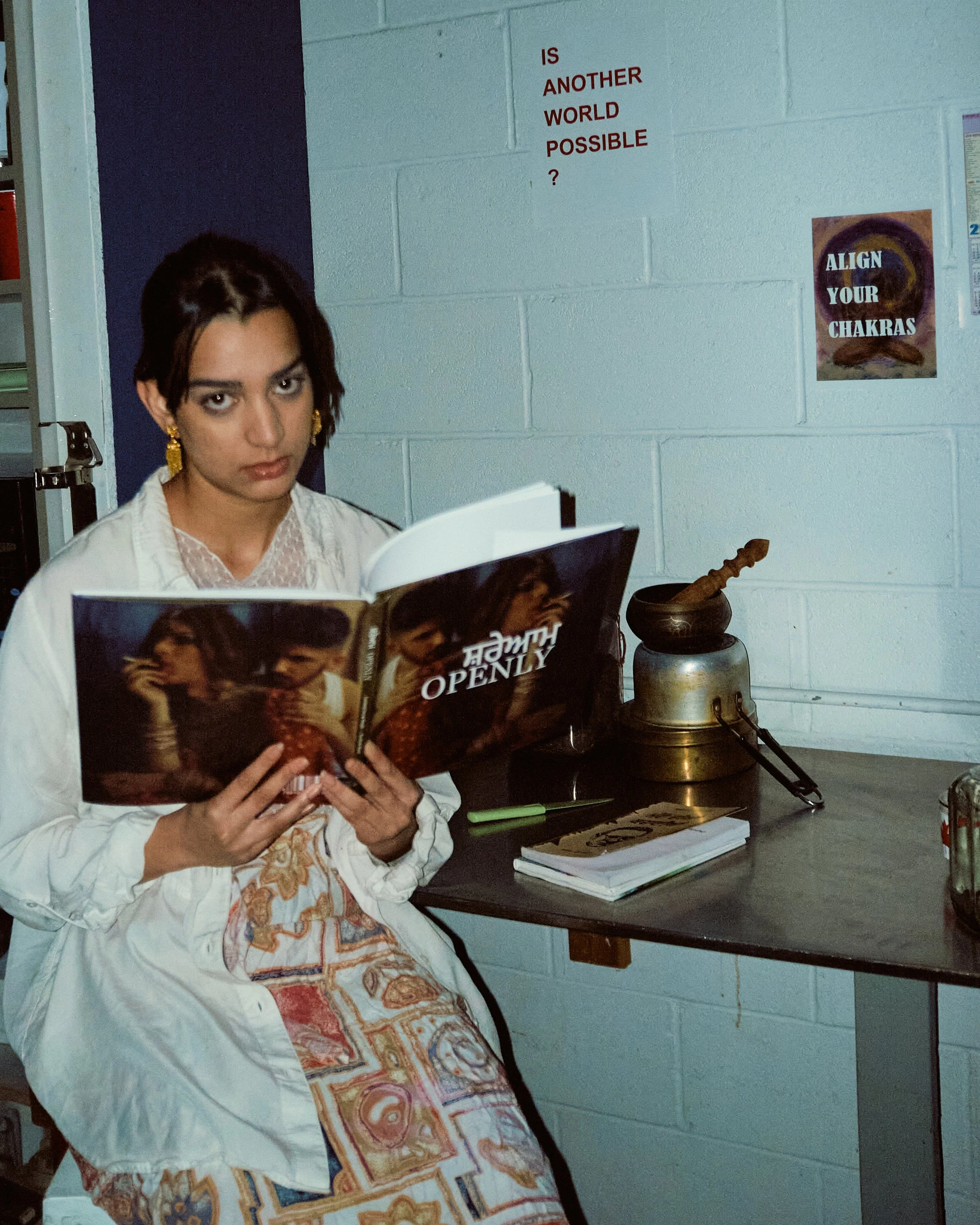 A woman with dark hair and gold earrings sitting at a desk, reading a book with a colorful cover, in a room with pale blue walls. Behind her, a sign on the wall says 'IS ANOTHER WORLD POSSIBLE?' and a poster that reads 'ALIGN YOUR CHAKRAS' with a spi