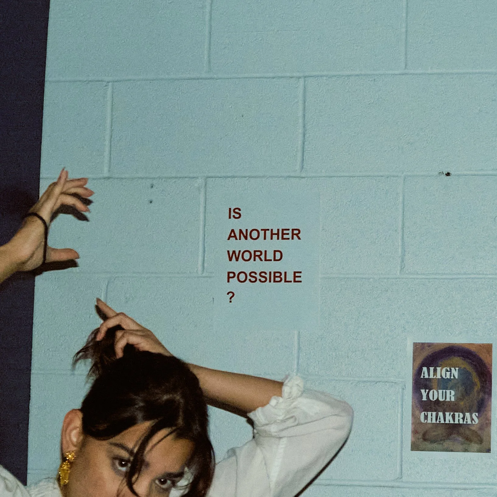A person with dark hair and earrings stretching their hand up near a turquoise painted cinder block wall with motivational posters and a sign reading "Is another world possible?" in red text.
