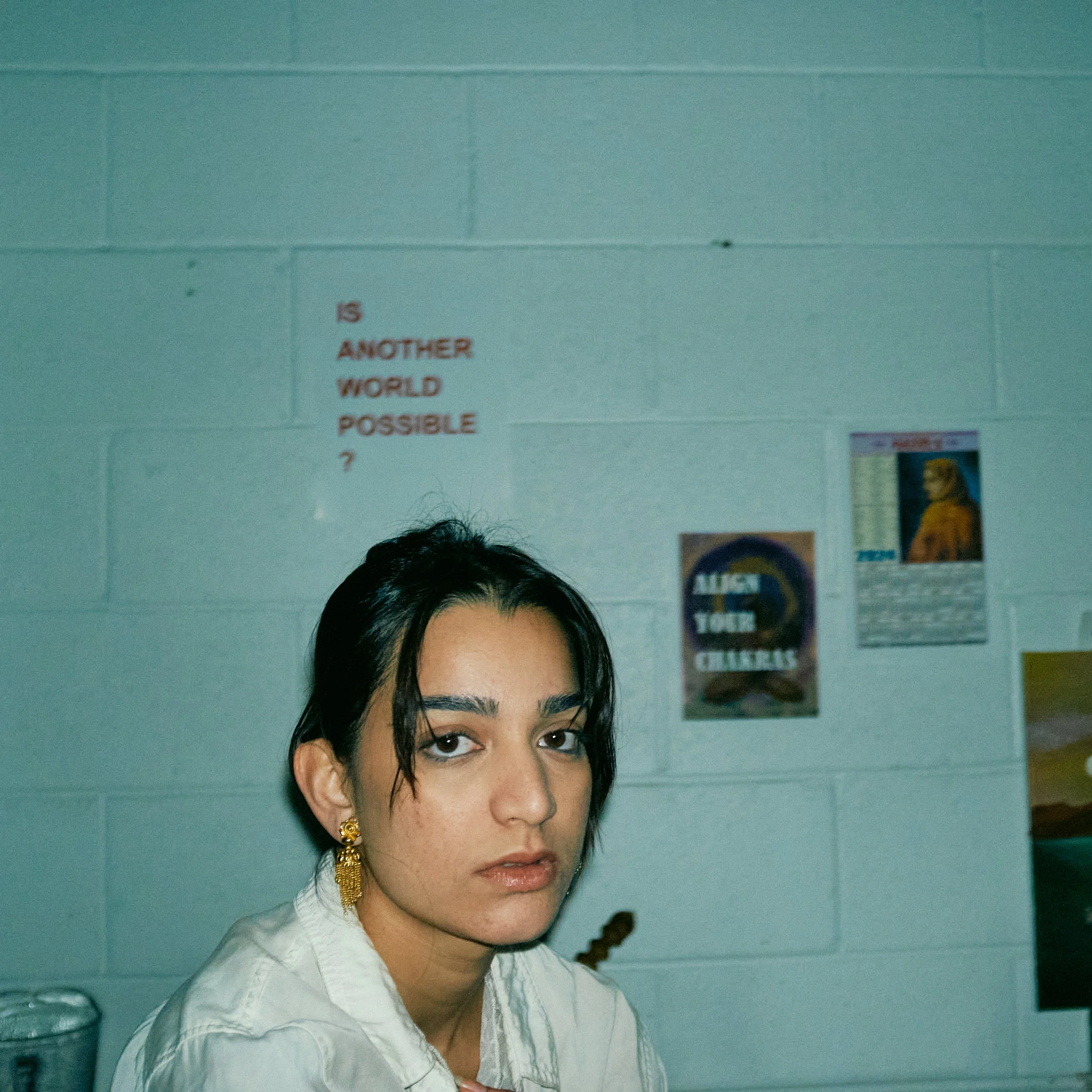 Portrait of a woman with dark hair, wearing gold earrings, and a white shirt, standing in front of a light blue wall with posters.