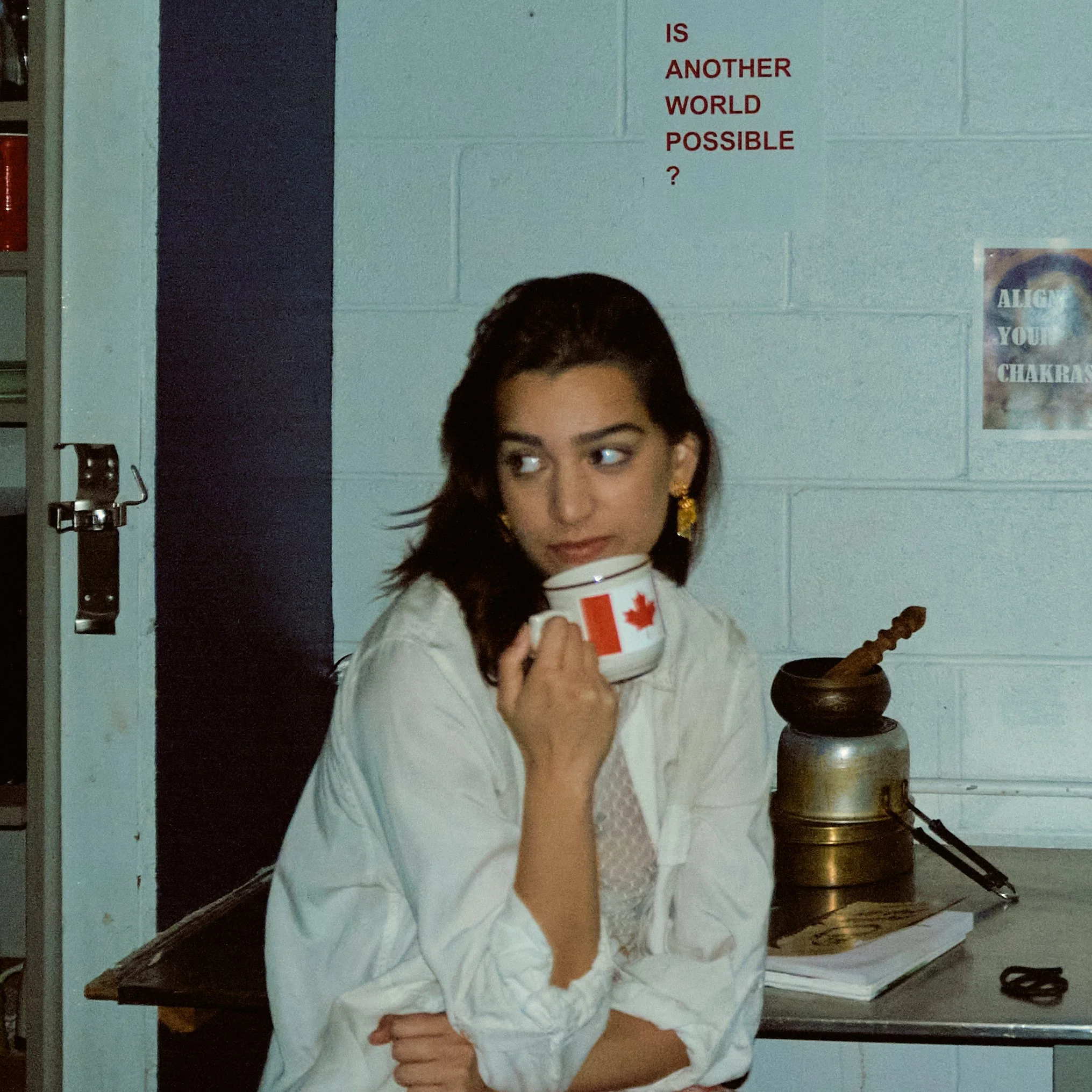 A young woman with dark hair and earrings, wearing a white top, holding a cup with a red maple leaf design, sitting at a table with a brass lamp on it, in a room with blue painted brick wall and posters.