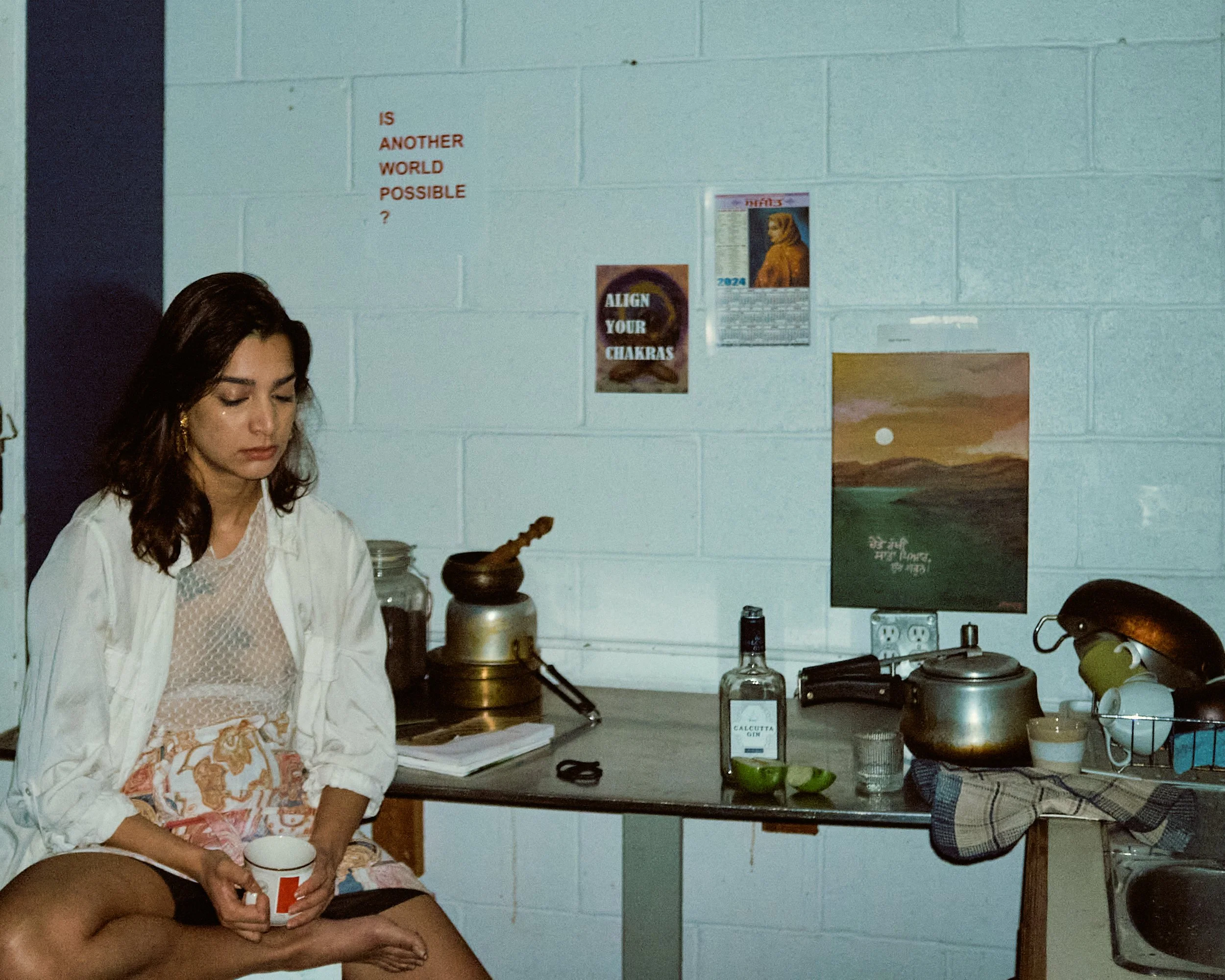 A woman with dark hair sitting on a kitchen counter, holding a white mug. The kitchen has light blue painted brick walls with various posters and a small painting on the wall. The countertop has kitchen utensils, a knife, and some small containers.
