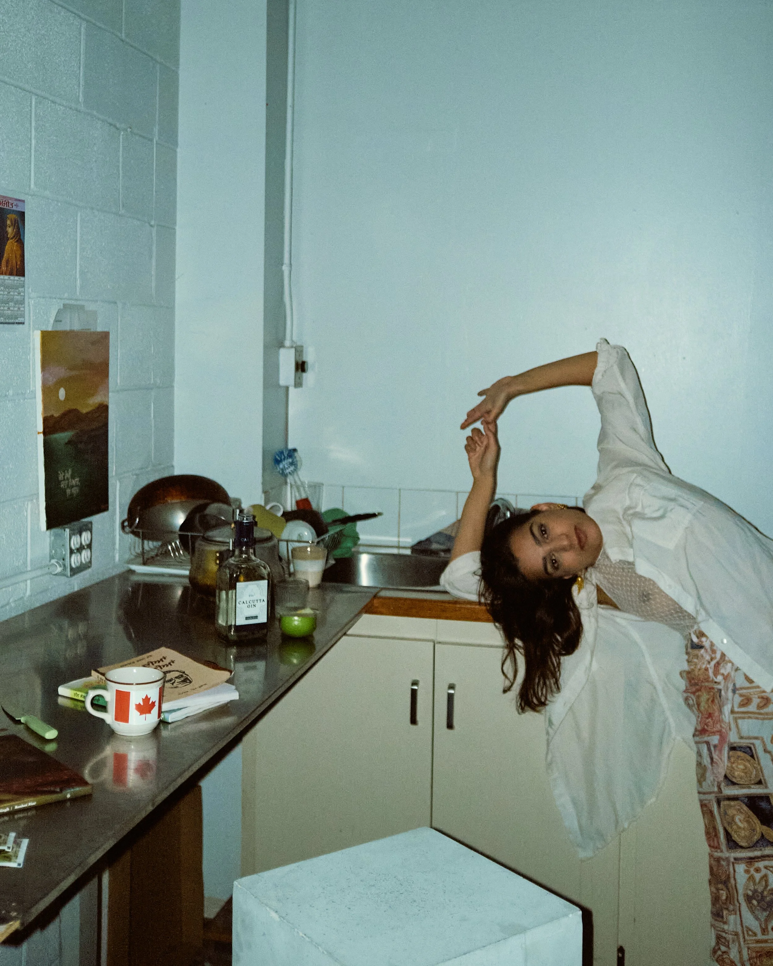 A young woman wearing a white dress with red patterns lying on a kitchen counter with her head resting on her hand, surrounded by kitchen items including a mug with a Canadian flag and a bottle of Calcutta gin.