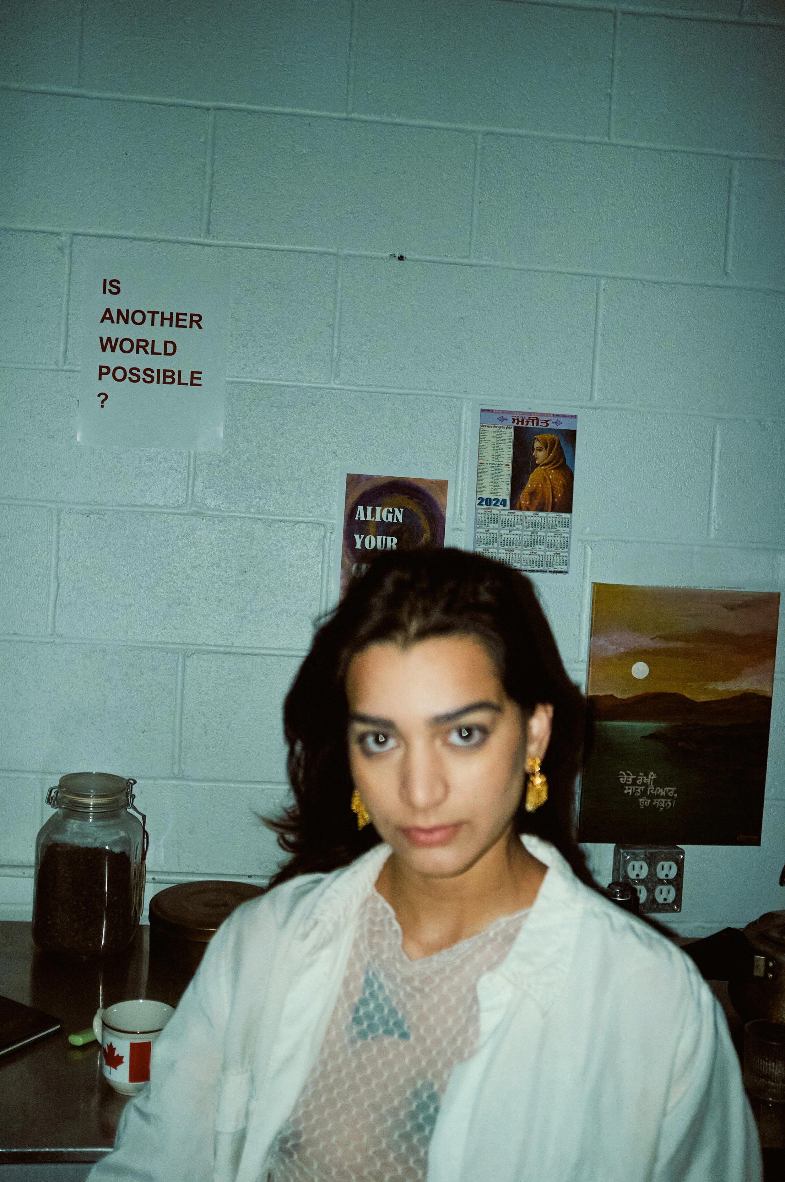A woman with dark, shoulder-length hair and gold earrings, wearing a white top with a lace pattern, is standing in front of a kitchen or workspace with a greenish cinder block wall. There are two jars and a mug on the surface in front of her, and pos