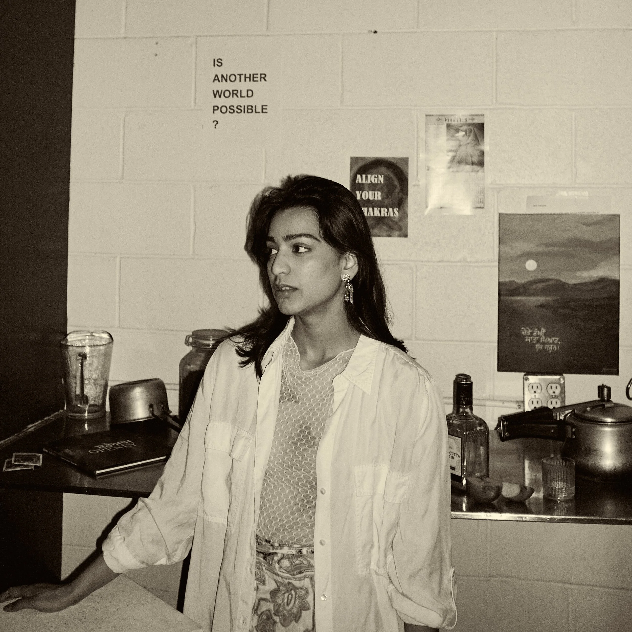 A person standing behind a table with various artwork and supplies, with posters and signs on the wall behind them. The person's face is partially visible.