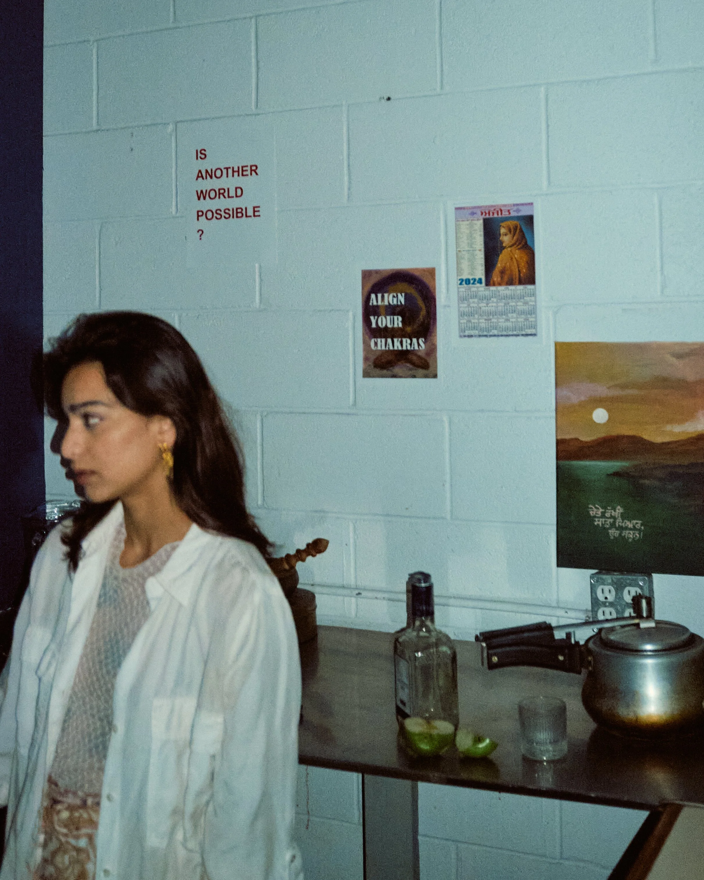 A woman with dark hair, earrings, and a white shirt, standing in a room with a gray brick wall. On the wall, several posters are visible, including a calendar, a message about chakras, and a poster with text partially visible. In the foreground, a ta