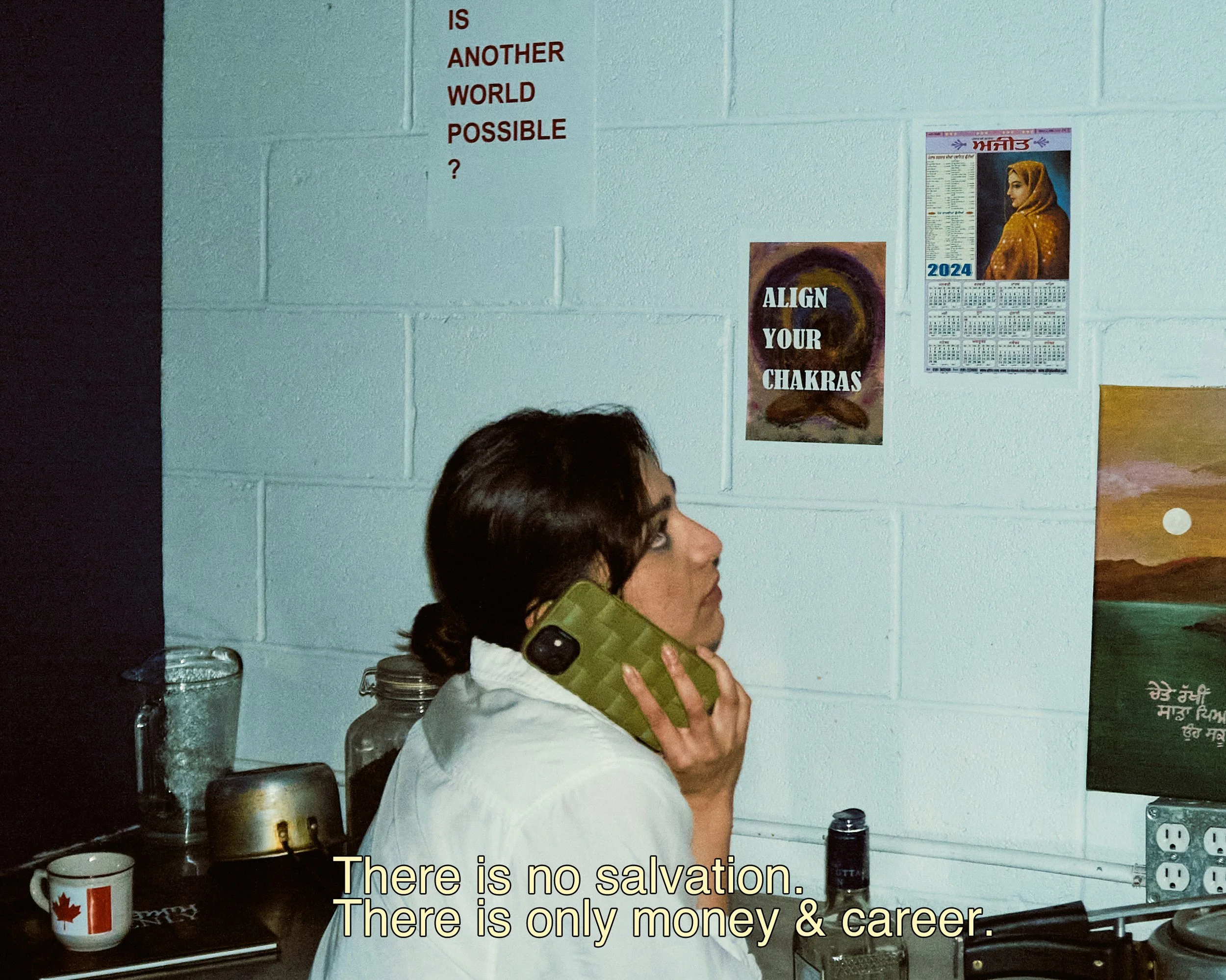 A woman wearing glasses talking on a green telephone in a kitchen. On the wall, there are motivational posters including one that reads 'ALIGN YOUR CHAKRAS' and another with the year 2024. The woman is dressed in a white hoodie, and there are various
