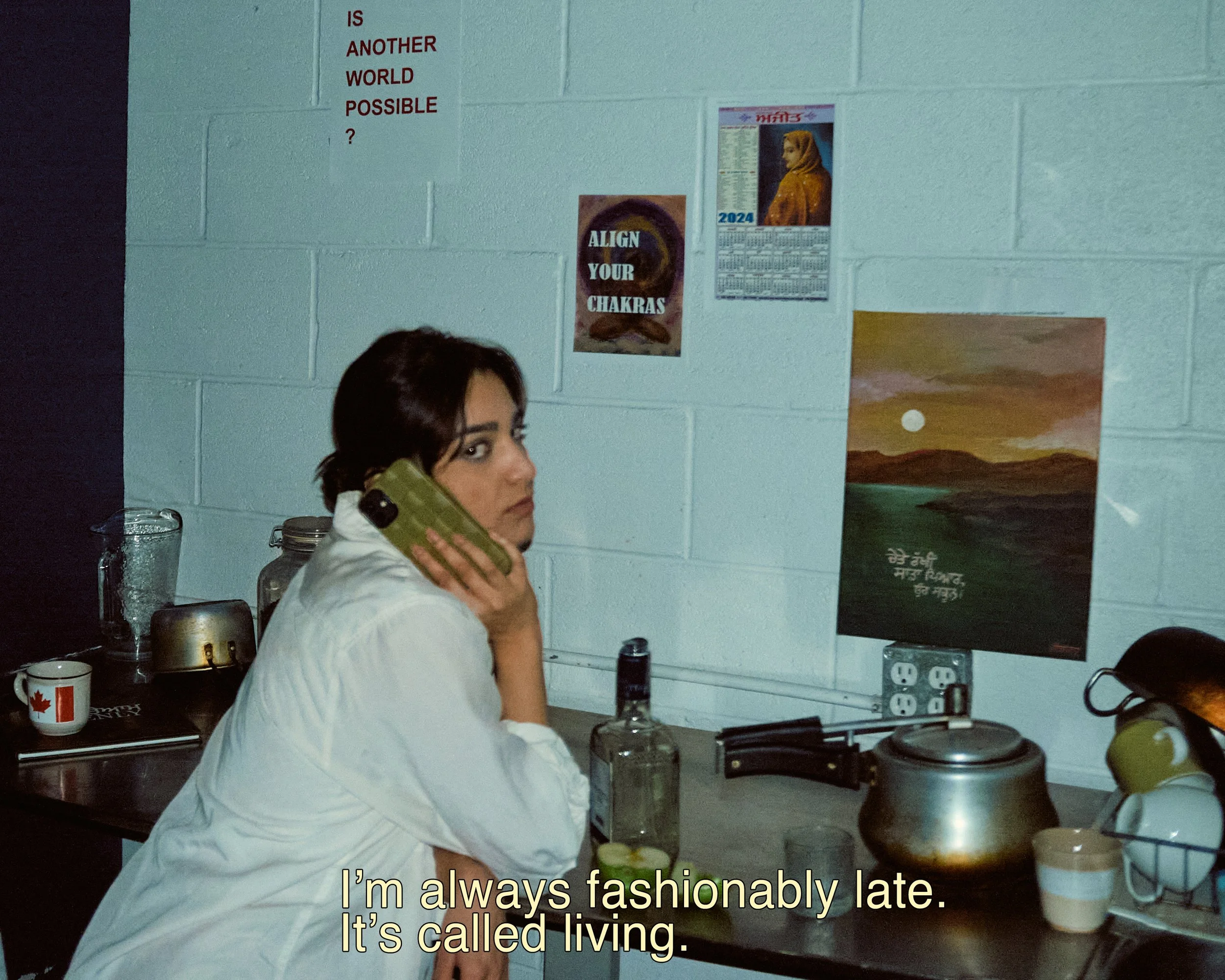 A young woman with dark hair wearing a white jacket speaks on the phone in a kitchen, with a backsplash, a calendar, posters on the wall, and various kitchen items on the counter.