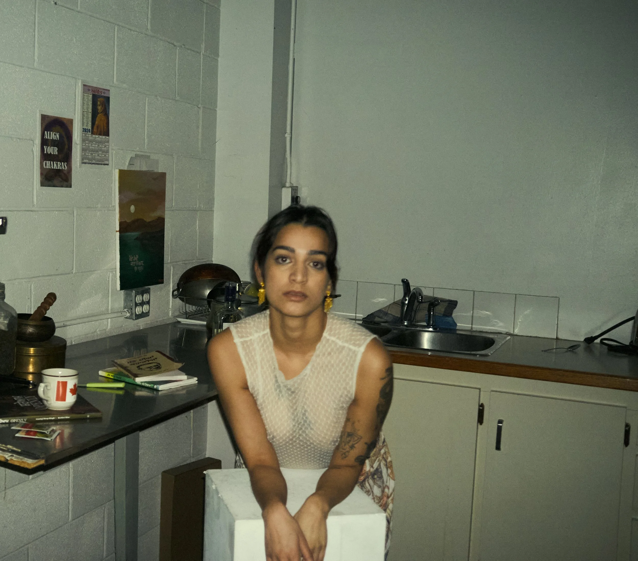 A woman with dark hair and tattoos on her arm standing in a kitchen with gray cinder block walls, a sink, and various kitchen items on the counter.