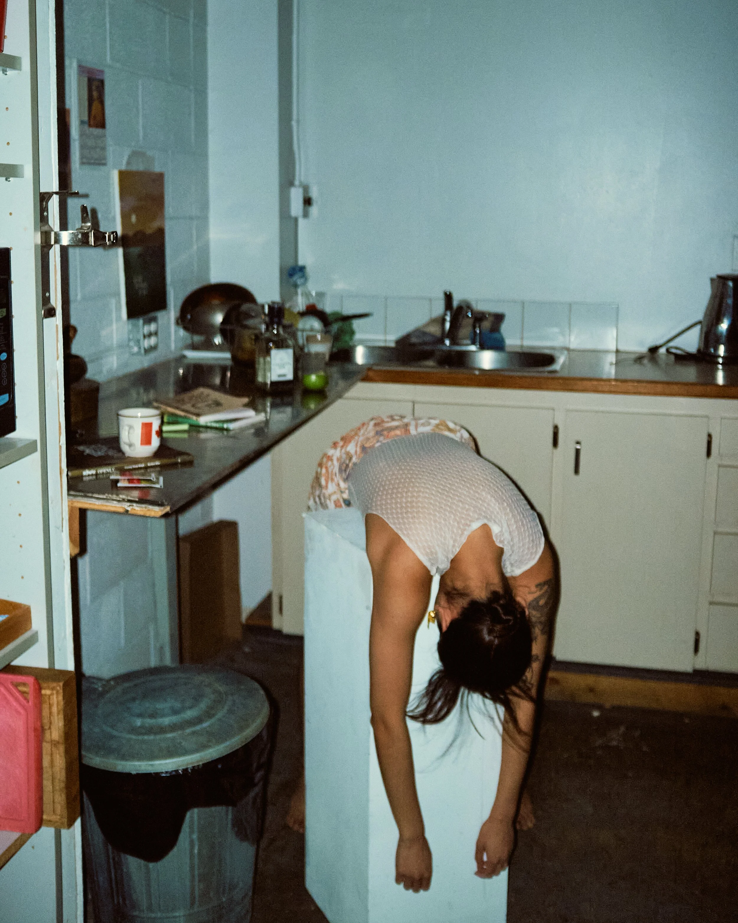 A woman with long dark hair bent forward, wearing a white dress with a floral scarf draped over her shoulders, in a kitchen with a countertop, sink, and various kitchen items.