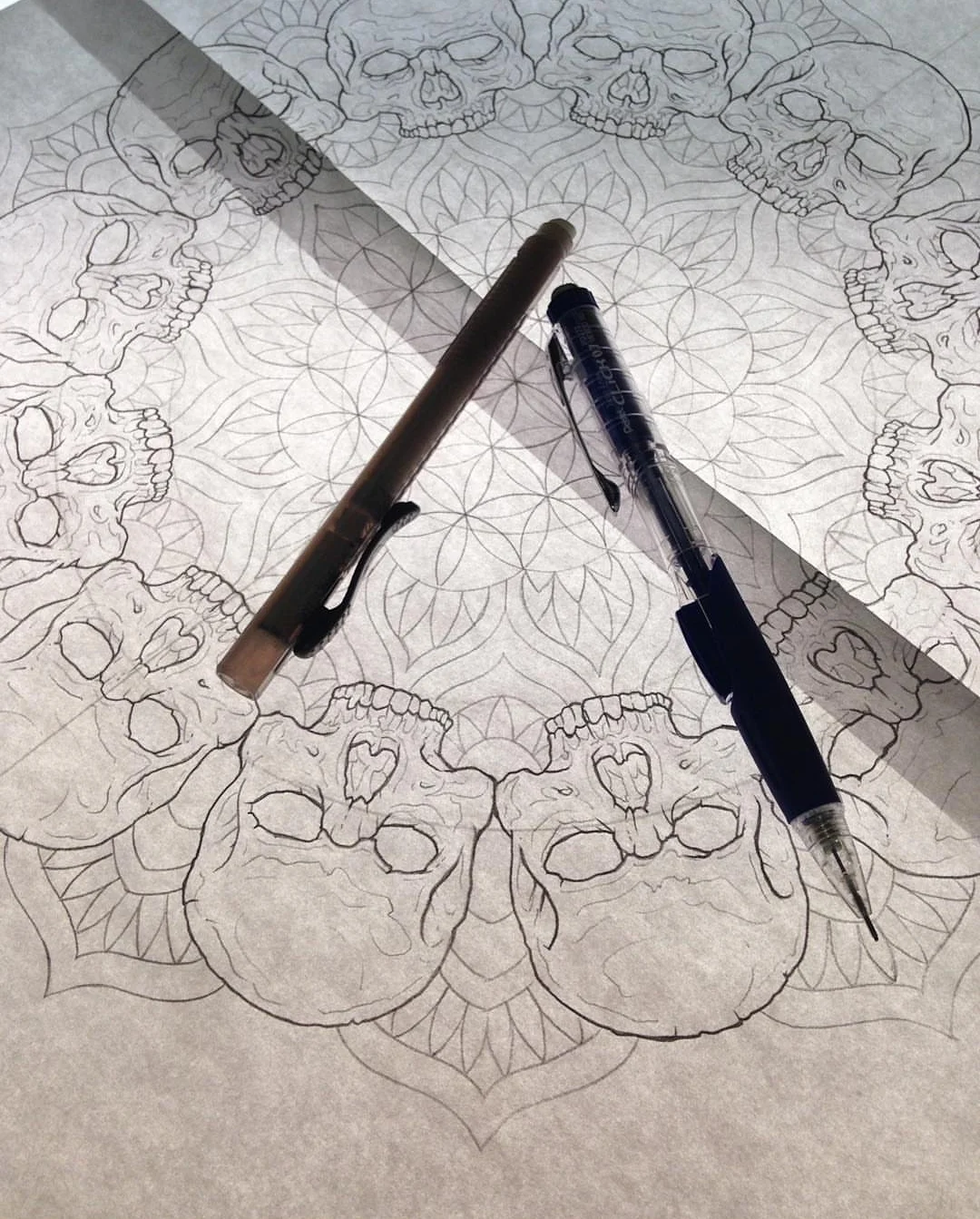 Drawing of skulls arranged in a mandala circle on patterned paper with two pens placed on top.