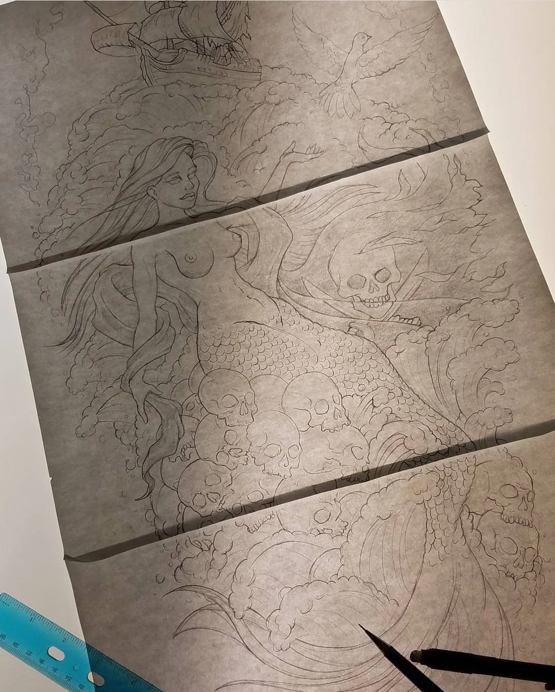 A detailed pencil sketch on tracing paper shows a mermaid with flowing hair and a scaly tail, surrounded by skulls and a large flaming skull with glowing eyes. In the background, there are waves, clouds, a ship in the sky, and a dove flying above.