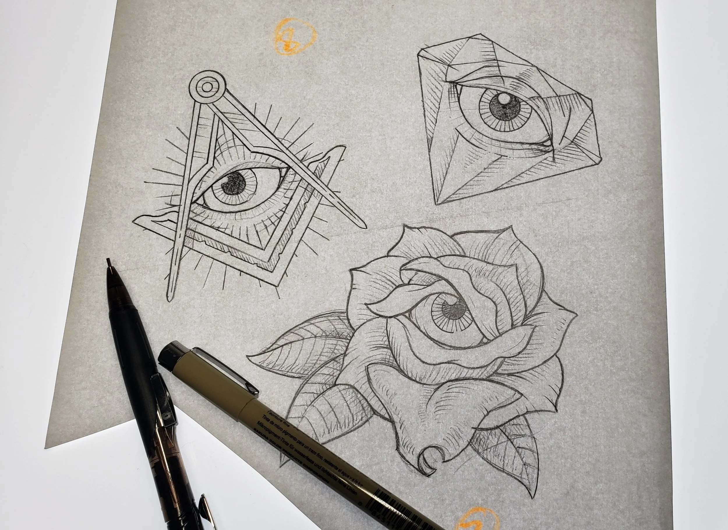 A sketch pad with detailed line drawings of eyes, a masonic square, a diamond with an eye inside, and a rose.