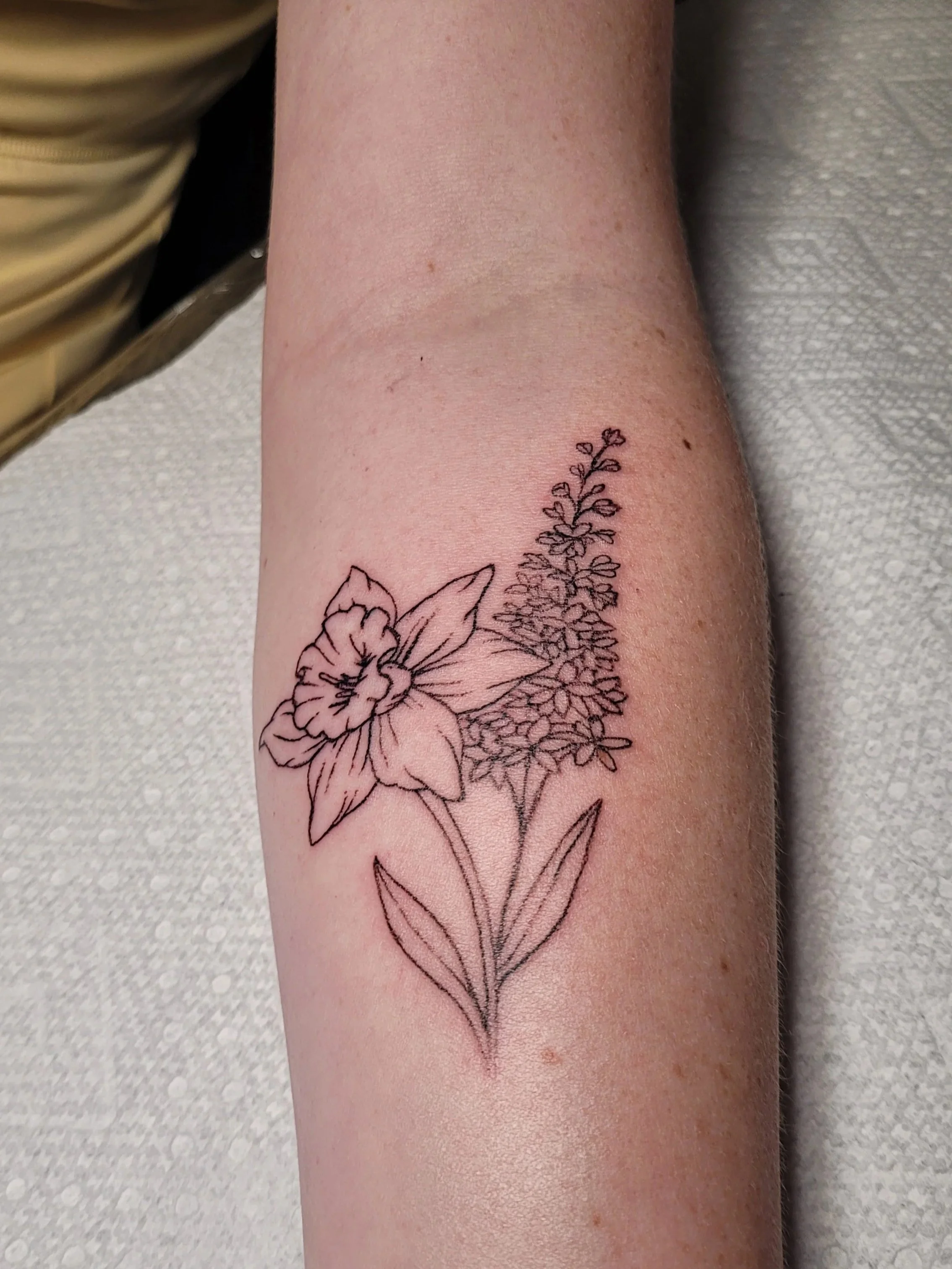 A person's arm with a black line tattoo of a floral design including a large flower, smaller flowers, and leaves.