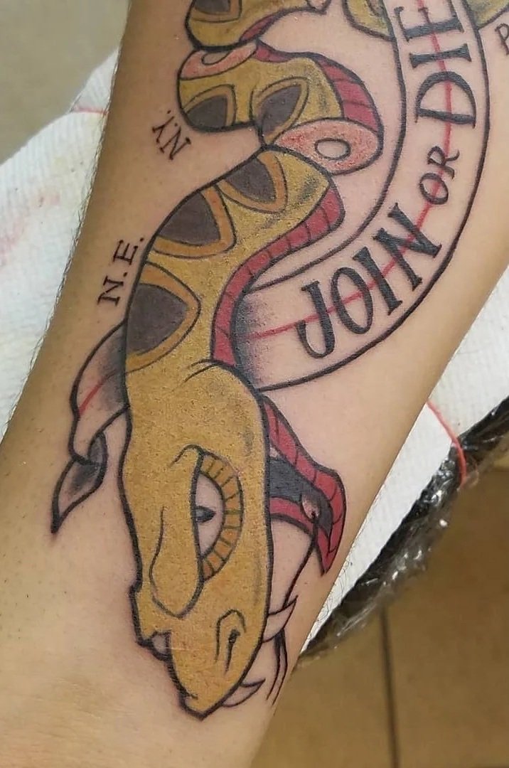 Tattoo of a yellow and brown snake coiled around a banner that reads "IN: E. JOR NO IDENT" with partial text visible.