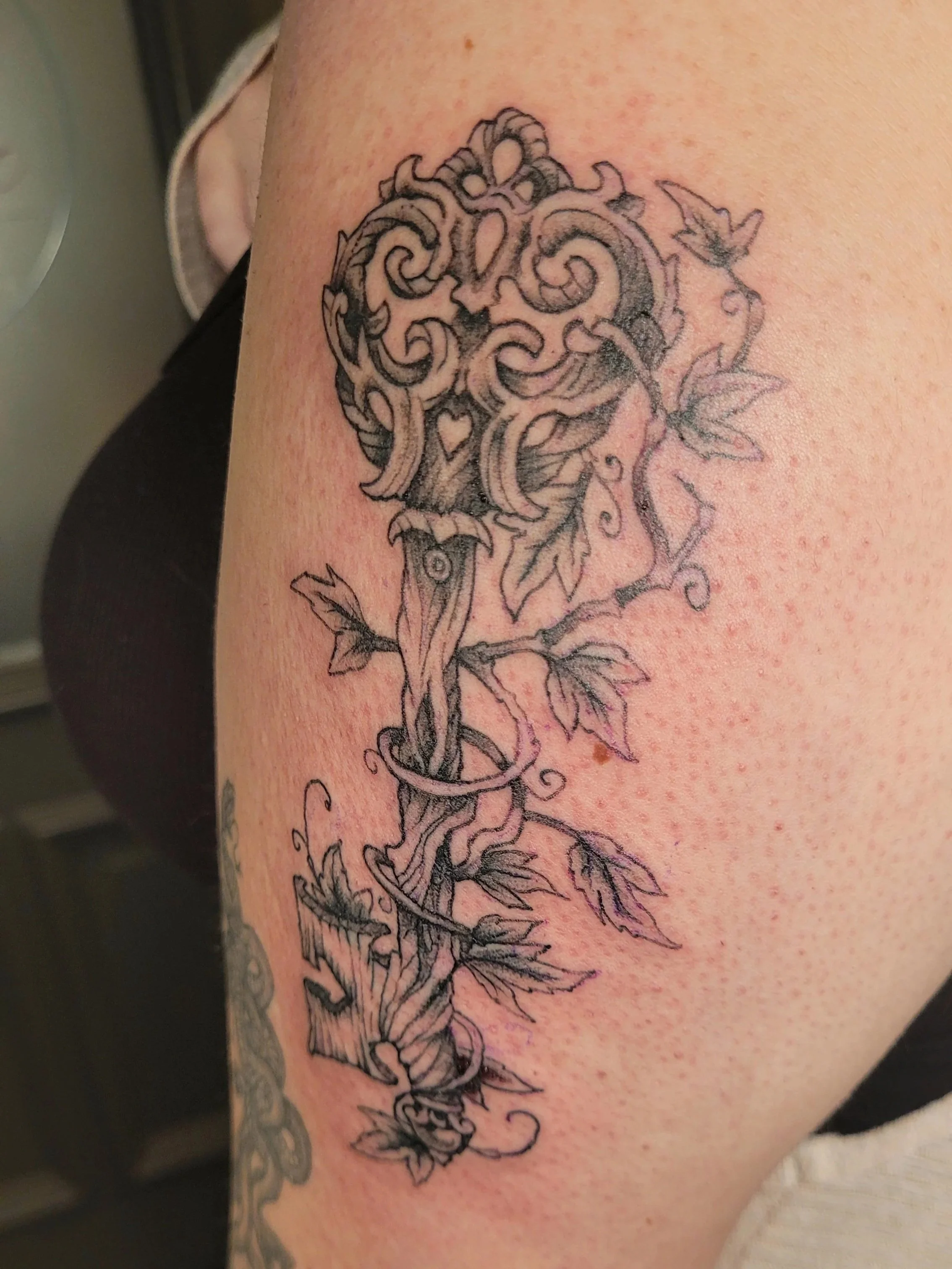 A tattoo featuring a stylized, intricate wooden key intertwined with floral elements, located on a person's upper arm.