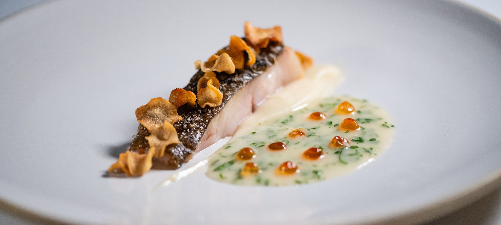 White fish topped with salsify chips beside a white and herb sauce with salmon roe